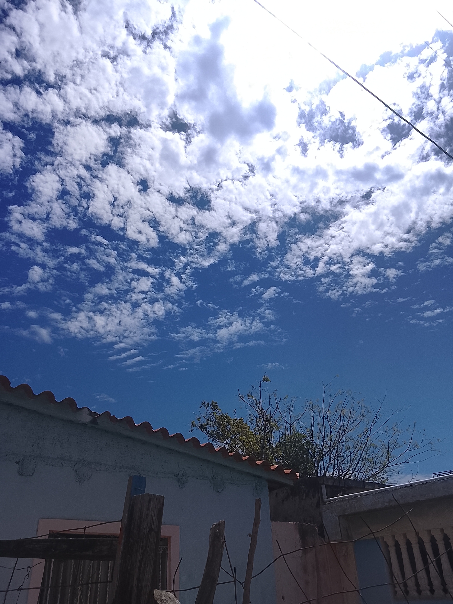 The image, taken in Altagracia, Venezuela, shows a bright blue sky filled with scattered white clouds. Power lines run across the upper right corner of the frame. In the foreground, the roofs of buildings with red clay tiles are visible. A fence and a tree with thin branches are also present. The buildings appear to be of modest construction.