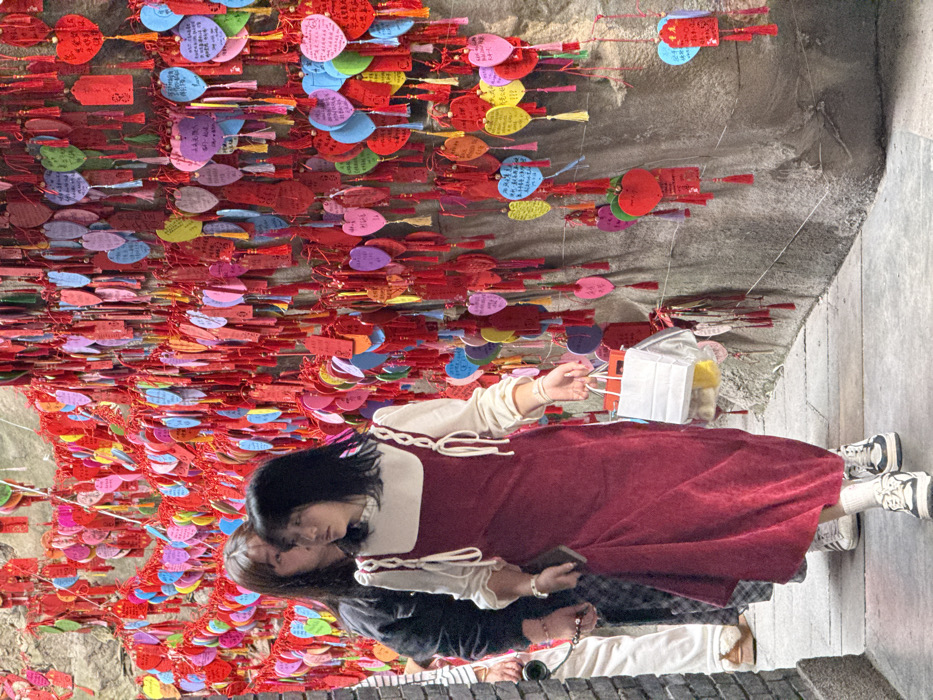 Two women stand amidst a dense array of colorful heart-shaped paper tags hanging from above. The woman in the foreground wears a long, burgundy dress over a cream-colored top. Her dark hair contrasts with her pale skin. She holds a small object, possibly a phone, in her hand. Behind her, another woman in a dark jacket leans towards her. Both are on a stone pathway. The background is filled with the multitude of tags, casting a vibrant, festive ambiance.