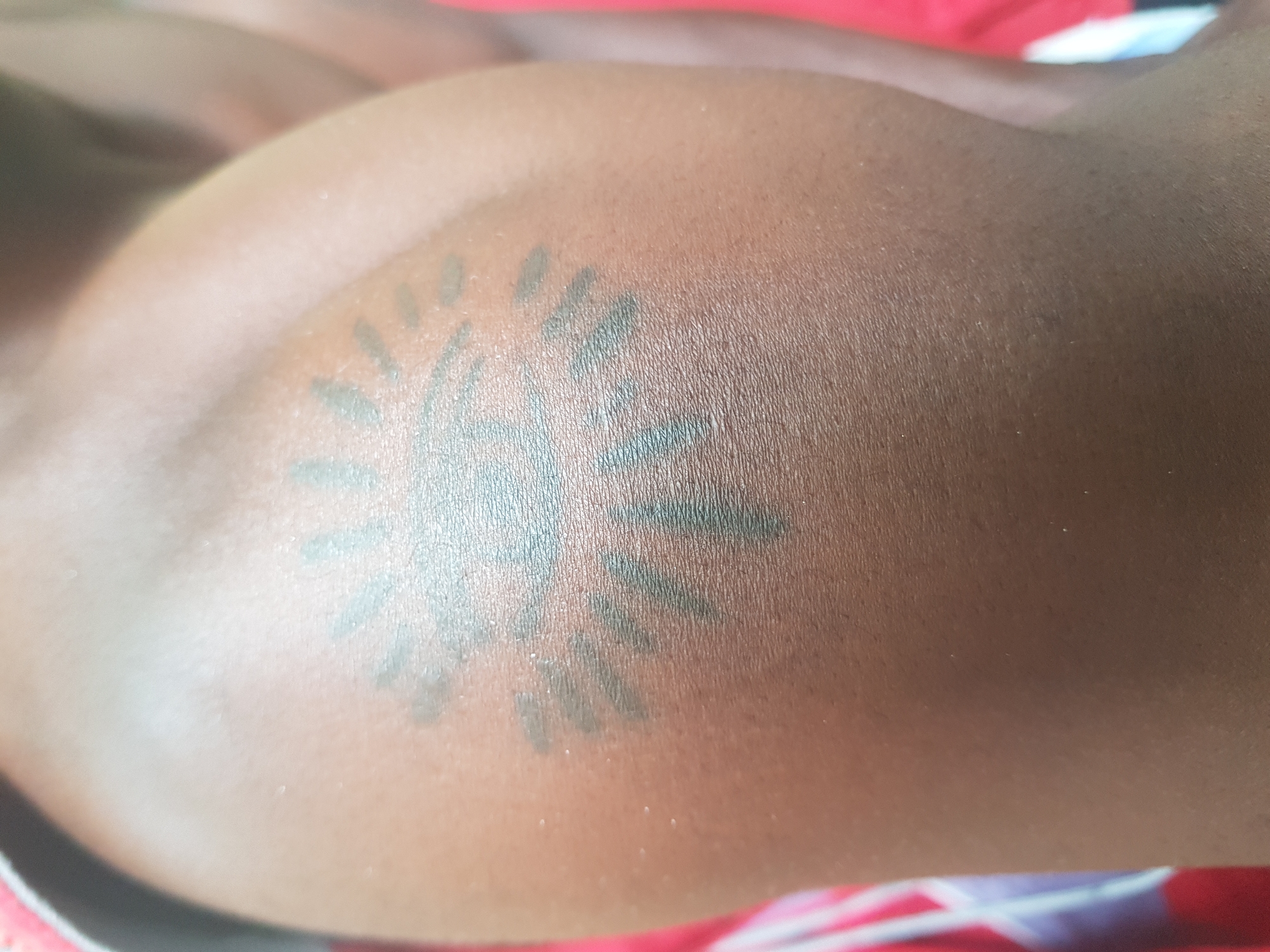 A close-up, medium shot shows a dark-skinned person's upper arm, likely the shoulder area, with a tattoo. The tattoo appears to be a dark ink design of an eye within a sun-like shape. The eye is depicted with a central pupil and surrounding iris-like markings. Rays radiate outwards from the central eye motif, forming the sun shape. The tattoo is positioned on the skin, which shows pores and texture. The background is slightly blurred, with hints of red and white fabric visible on the left side, and a lighter-skinned area on the right, suggesting another body part or clothing. The lighting is moderate, creating some shadows and highlighting the skin's surface.