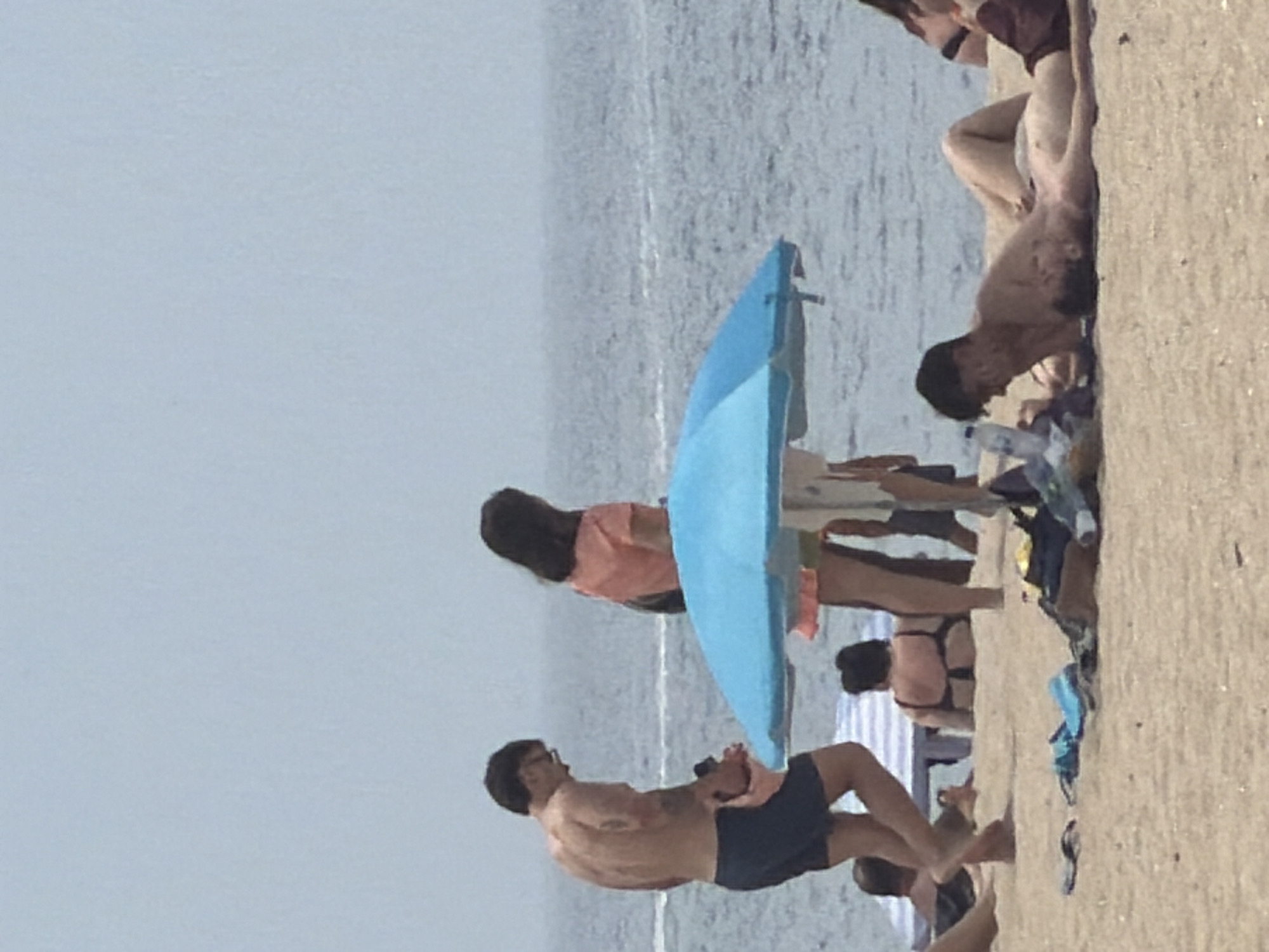 The image depicts a beach scene on a slightly overcast day, with the ocean in the background. In the foreground, a man in dark blue swim trunks stands facing left, his back to the viewer, looking downwards. To his right, a woman in an orange top and a light-colored skirt stands, facing away from the camera. A blue beach umbrella is prominent in the middle ground, partially obscuring a beach chair and another person lying down. Further back, a woman in a black bikini is seated on the sand. The beach itself is sandy, with various items of clothing and a water bottle scattered on it. The scene is likely the Praia da Baía in Espinho, Portugal, a popular urban beach known for its wide sandy expanse and proximity to the city.