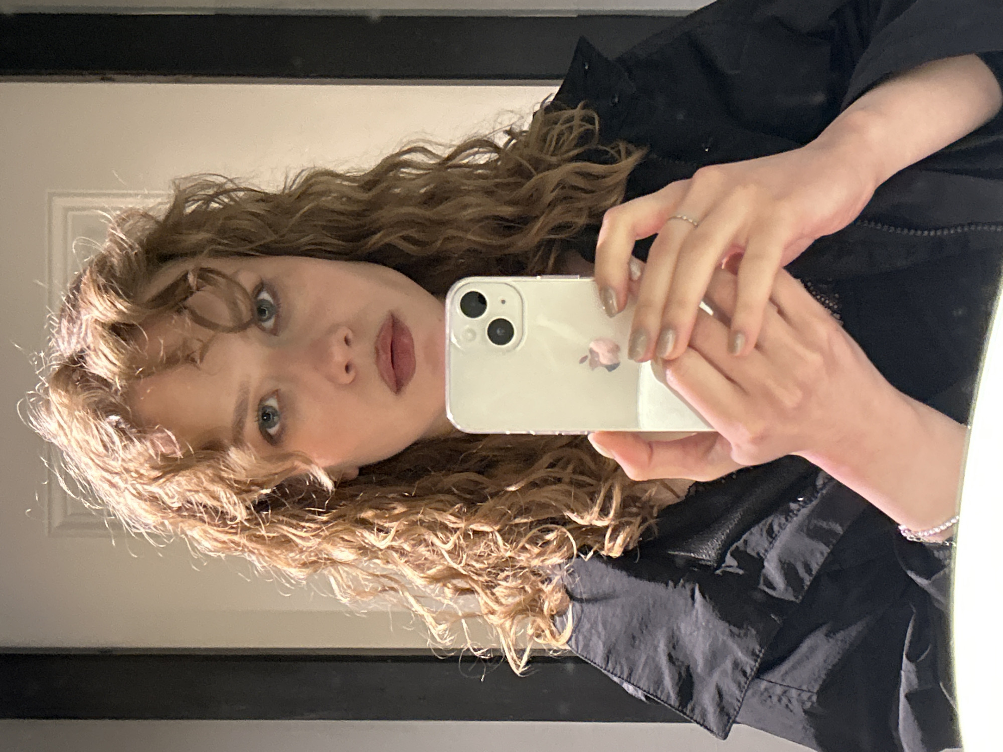 This is a medium shot, indoor, selfie-style photograph of a young woman with curly, light brown hair. She is holding a white smartphone in her hands, with the screen facing her to capture the reflection. The woman is wearing a black jacket with a high collar. Her left hand, visible in the foreground, has a thin ring on one finger and a sparkling bracelet on her wrist. Her right hand is partially obscured by the phone. The background appears to be a bathroom or dressing area, with a white wall and a dark vertical element, possibly a door frame or mirror. There is a subtle reflection of the woman in the phone's screen. The image is well-lit, with a soft light source. There is no identifiable landmark or context to suggest a location in Saint Petersburg, Russia.