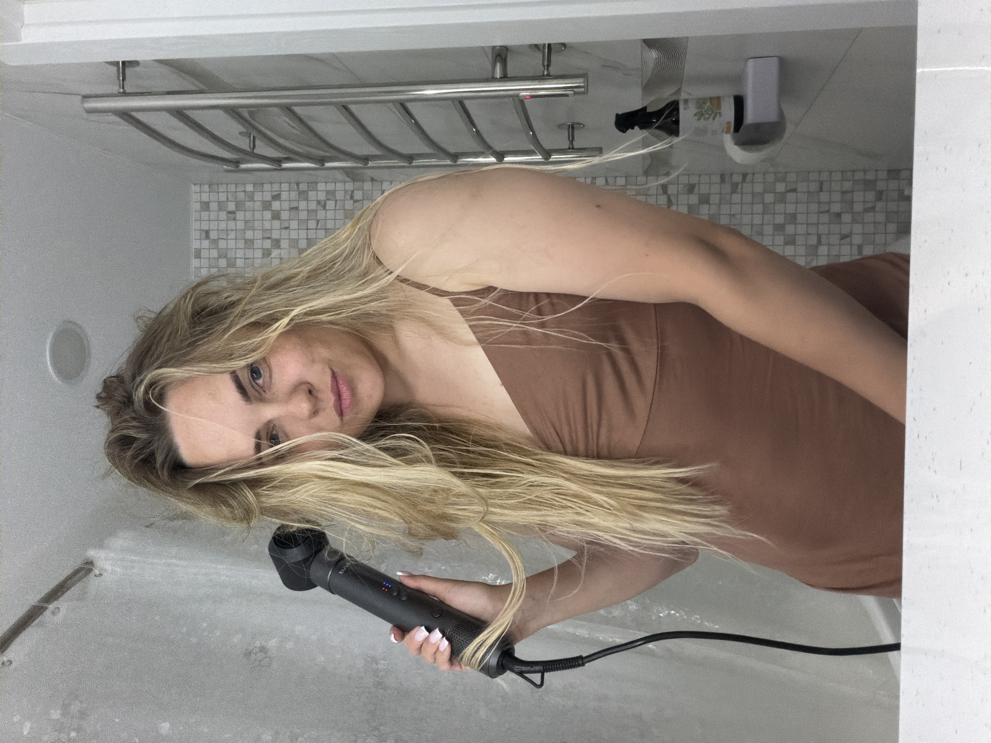 A woman with long, blonde hair is holding a hairdryer in a bathroom. The bathroom has a shower curtain, a towel rack, and tiled walls. The woman is wearing a brown top.
It is not possible to determine the location in the city of Votkinsk, Russia.