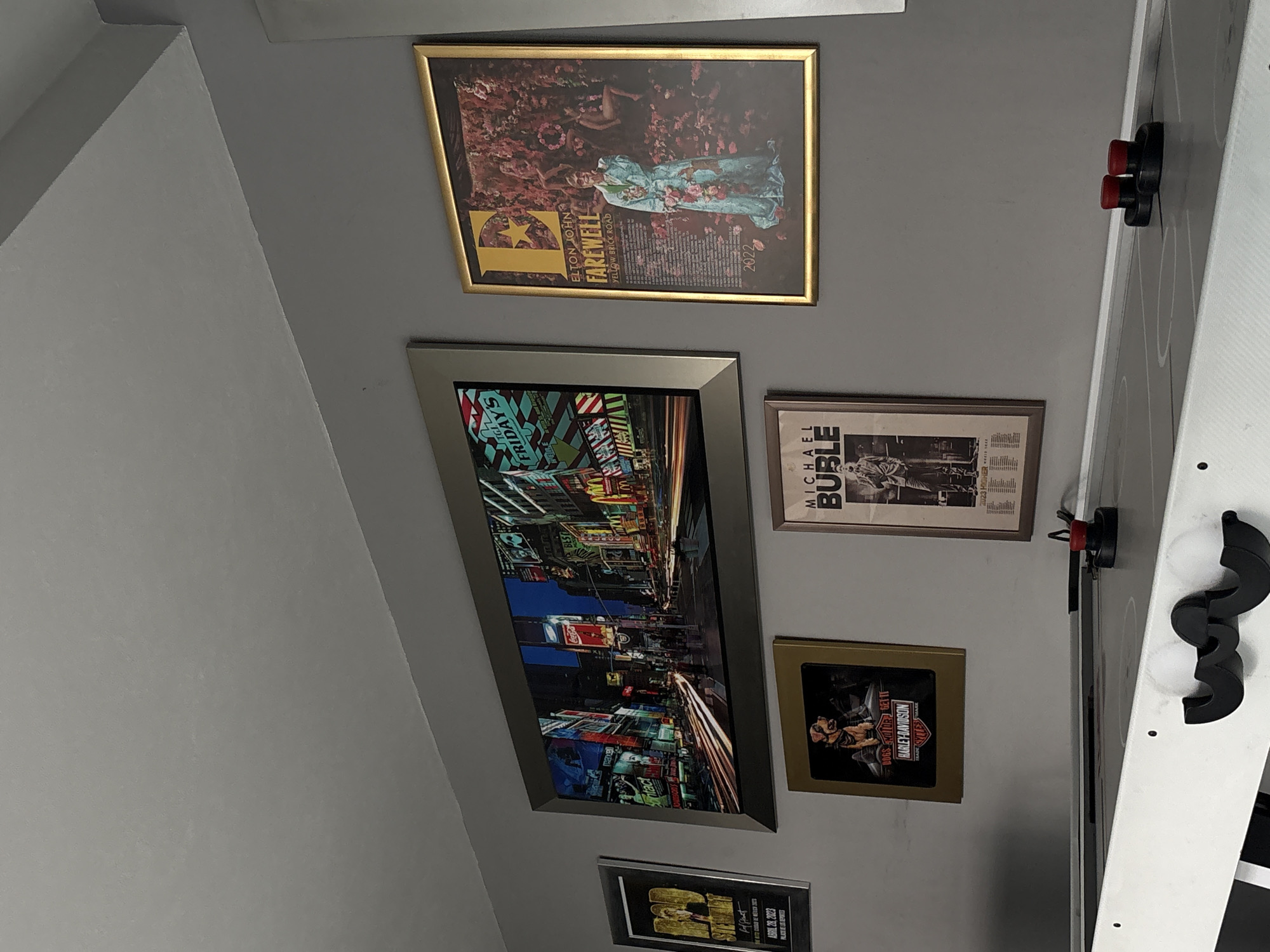 The image shows a wall with a collection of framed posters and pictures.
Starting from the top left and moving downwards, there's a poster with a gold frame featuring a person in a blue floral dress, identified as Elton John's "Farewell Yellow Brick Road" tour from 2022.
Below it is a larger, horizontally oriented picture in a silver frame that depicts a cityscape with bright lights and numerous signs, one of which reads "FRIDAY'S".
To the right of the cityscape poster is a framed poster of Michael Buble and below the frame is a poster for "Harley-Davidson" with a gold frame, next to it.
Further down is a framed certificate of Abril 28, 2023.
Along the right side of the frame, there is part of what appears to be a white object,possibly an air hockey table with some black accessories or pieces visible.
The wall is painted in a neutral gray color, and the ceiling is visible on the top left.