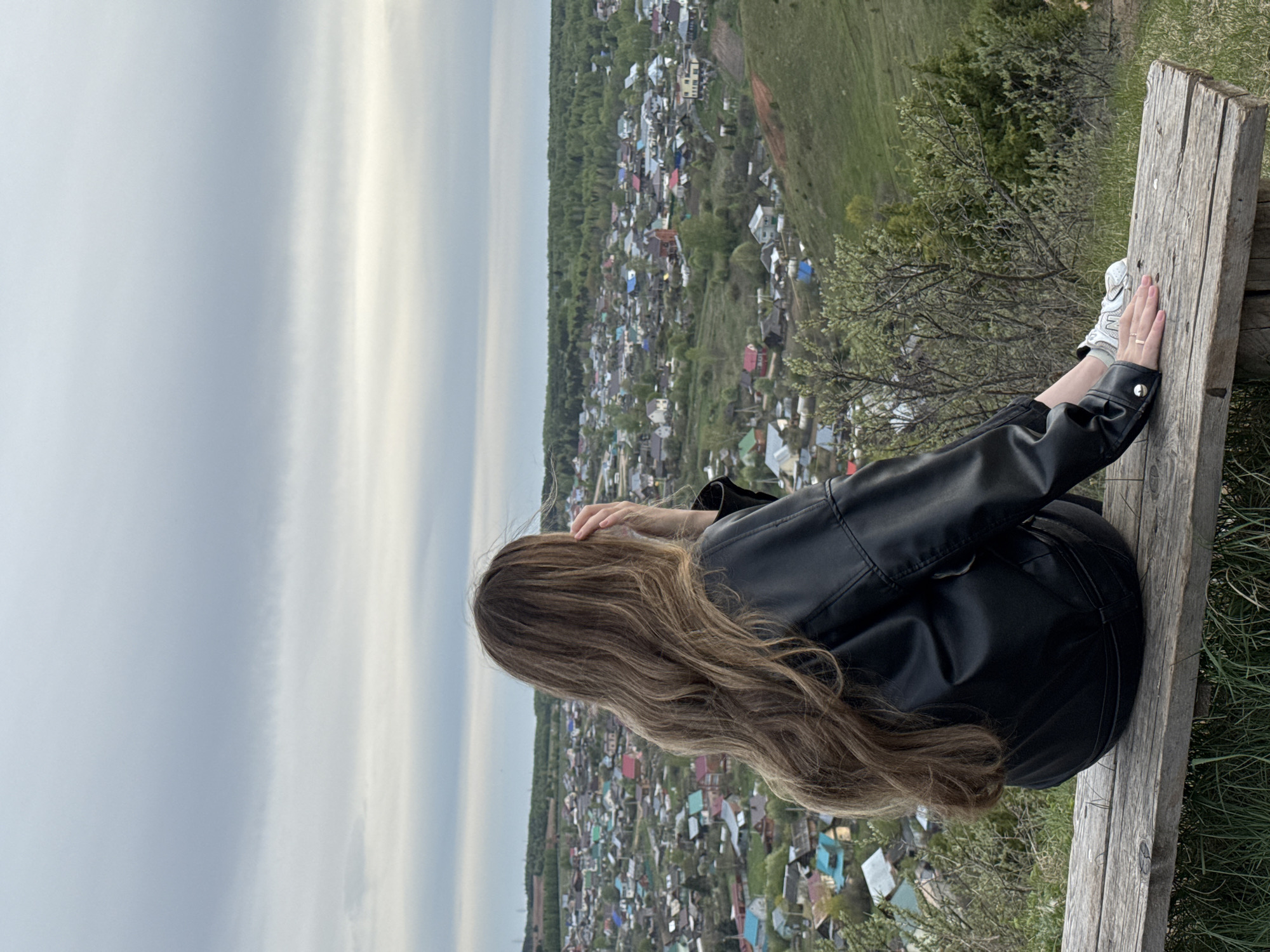 A young woman sits on a wooden bench overlooking the city of Ольховка, Russia. The woman wears a black leather jacket and black pants. Her long, light brown hair cascades down her back. She rests her hands on her head. The bench is made of rough-hewn wood. The city below consists of a dense collection of small houses with colorful roofs, interspersed with trees. The sky is overcast.