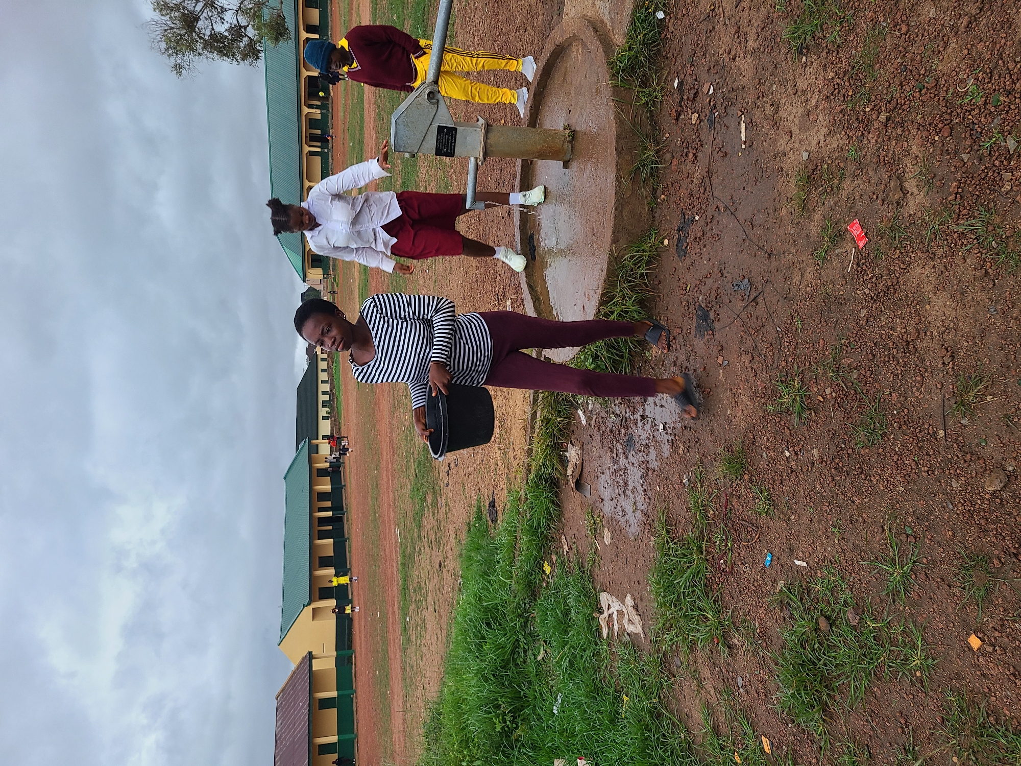 The image, set in Jos, Nigeria, captures three young women gathered around a hand pump outdoors, likely within a school or community compound. In the foreground, a woman wearing a black and white striped long-sleeve top and dark leggings holds a black bucket, looking towards the viewer with a slight smile. Behind her, another woman in a white long-sleeve shirt and dark red shorts operates the grey metal hand pump, from which water appears to be flowing, creating wet, muddy patches on the reddish-brown ground around its concrete base. A third person, dressed in a maroon long-sleeve top and yellow track pants, stands further back to the right of the pump.
The ground consists of reddish soil interspersed with patches of green grass, and some litter, including a red wrapper, is visible. In the background, several long, single-story school buildings with green roofs and beige or yellow walls are visible under an overcast sky, suggesting a cloudy daytime. A small, illegible plaque is affixed to the side of the hand pump. The scene depicts the daily activity of fetching water, characteristic of reliance on boreholes in the region.