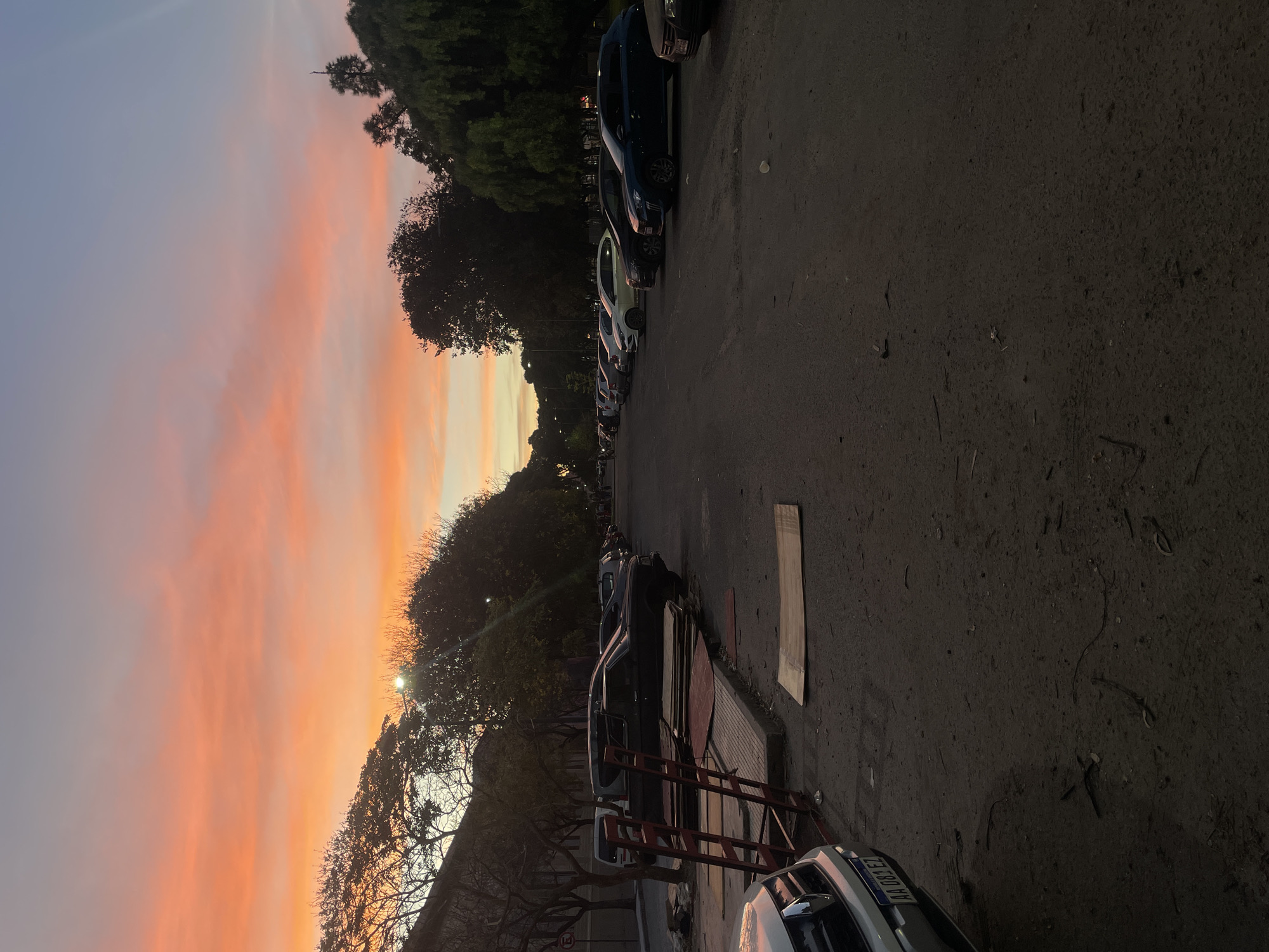 The image shows a street scene in Buenos Aires, Argentina at sunset. Cars are parked along the right side of a paved street, which takes up most of the foreground. On the left side, there are trees silhouetted against a vibrant orange and pink sky. A streetlight is visible near the base of the trees. In the bottom right corner, a vehicle is visible with a license plate. There are also metal barriers and construction materials like wooden sheets along the sidewalk. The overall scene is calm and quiet, with the colorful sunset being the most striking feature.
