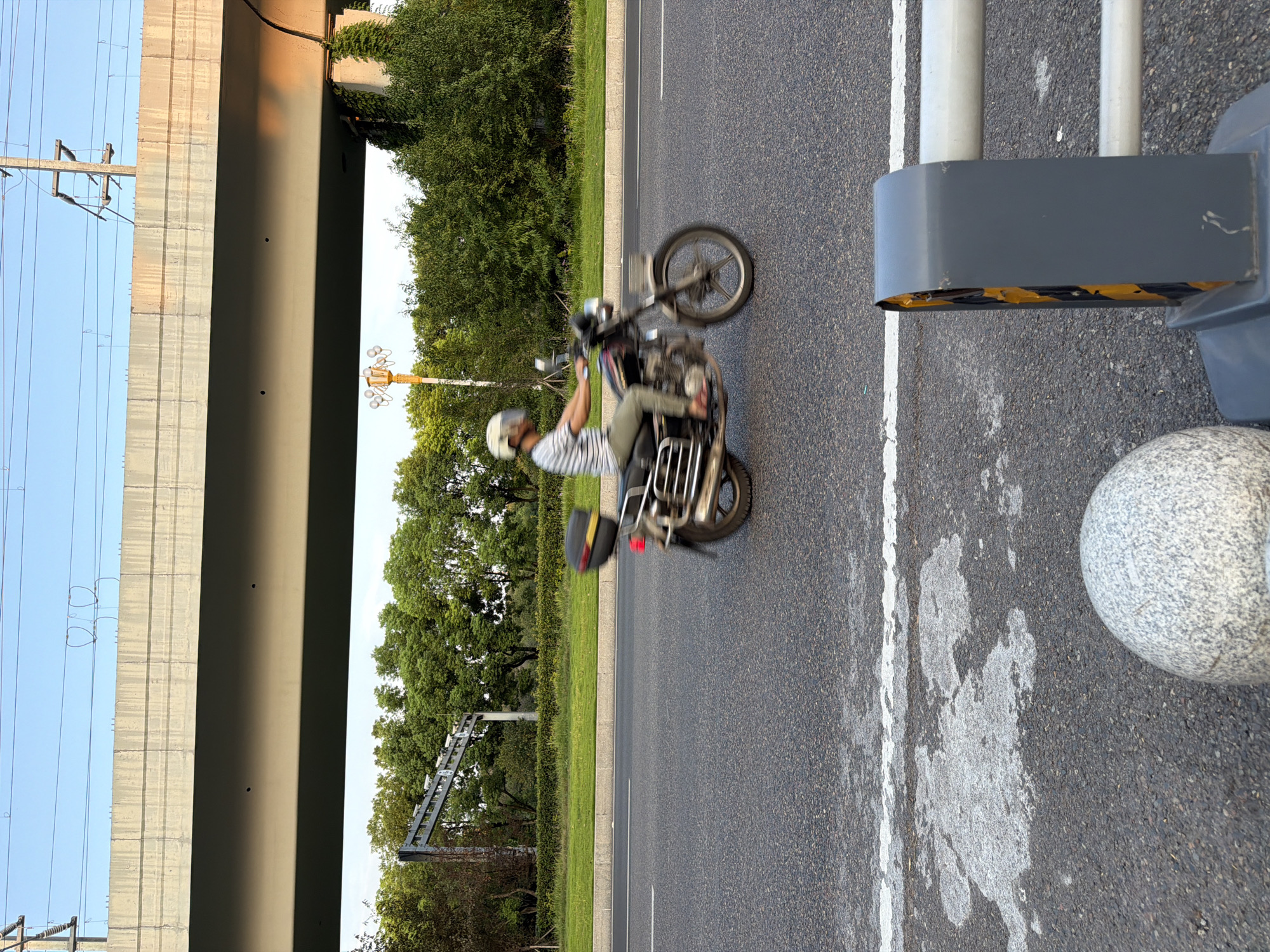 A side view captures a motorcyclist in motion on a paved road under a concrete overpass. The motorcyclist, wearing a helmet and a striped shirt, is moving from left to right. The motorcycle has a black storage box on the back. Green trees and foliage line the side of the road, with a utility pole and streetlights visible in the background. The sky is clear and blue, indicating daytime. The scene appears to be in an urban or suburban area, possibly in Jiangjin, China, given the presence of overpasses and the overall aesthetic.
