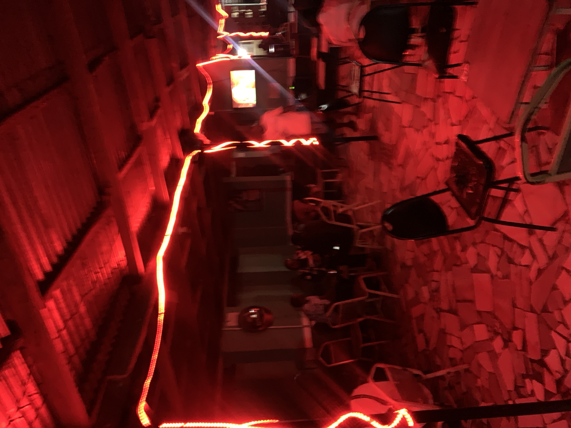 The image depicts an indoor area in New Stadium, Nigeria, possibly a restaurant or bar, illuminated with intense red lighting. Strings of red LED lights are strung along the ceiling and walls. The floor appears to be tiled with irregularly shaped stones or bricks. Several chairs are visible, some made of metal and others of dark plastic. Some people are seated at tables in the central part of the room. Overall, the environment seems dimly lit and the red lighting gives it an unusual cast.
