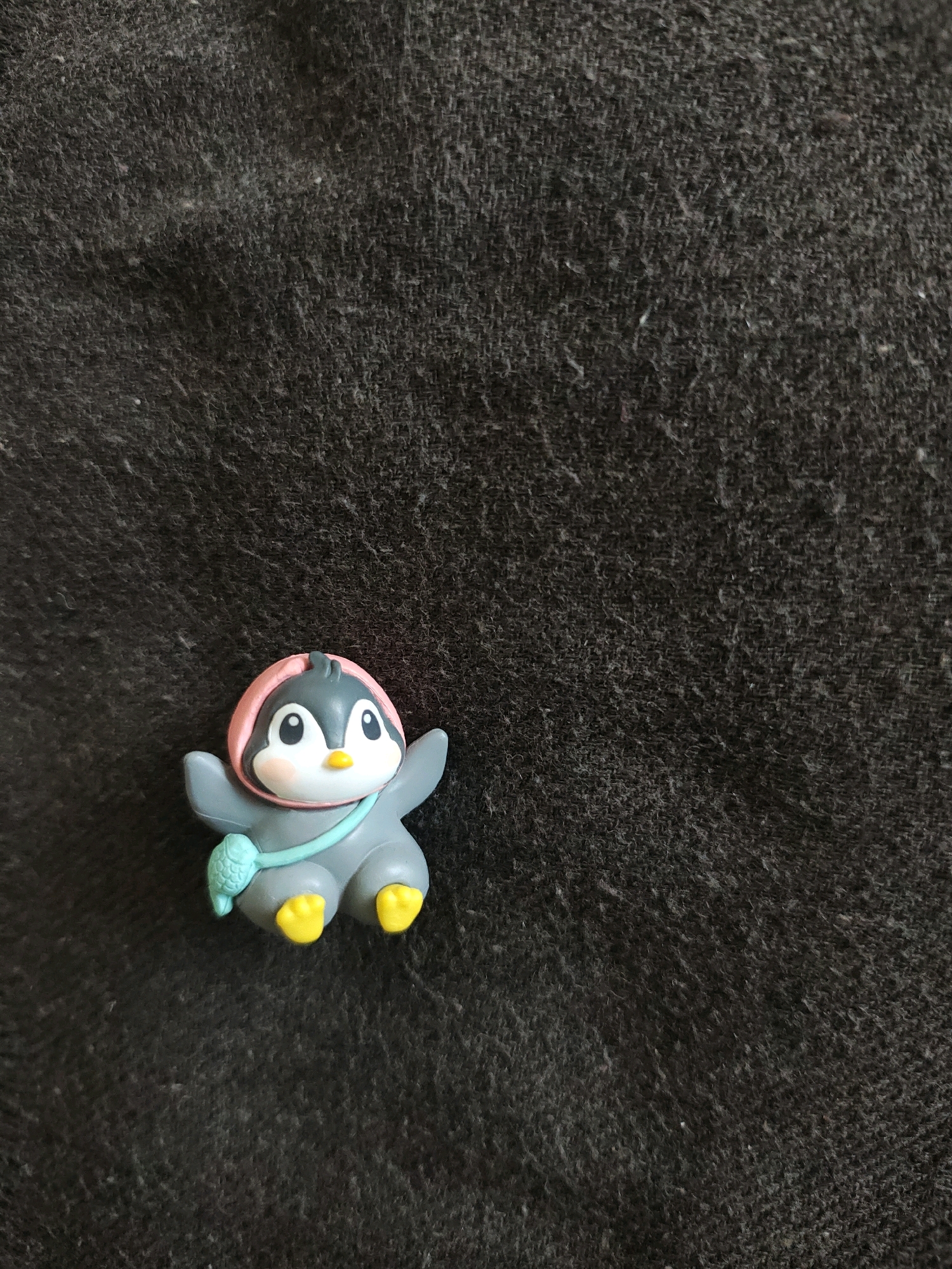 A small, gray penguin figurine with yellow feet and a pink hood is positioned on a dark, textured surface. The figurine has large, white eyes with black pupils, pink cheeks, and a yellow beak. A light blue string is draped around its neck, with a small, green, fish-shaped charm attached to it. The surface appears to be fabric, possibly a blanket or upholstery, with a visible weave and some lint or dust particles. The lighting is neutral, casting minimal shadows. This scene is not located in the city of Jaipur Municipal Corporation, India.