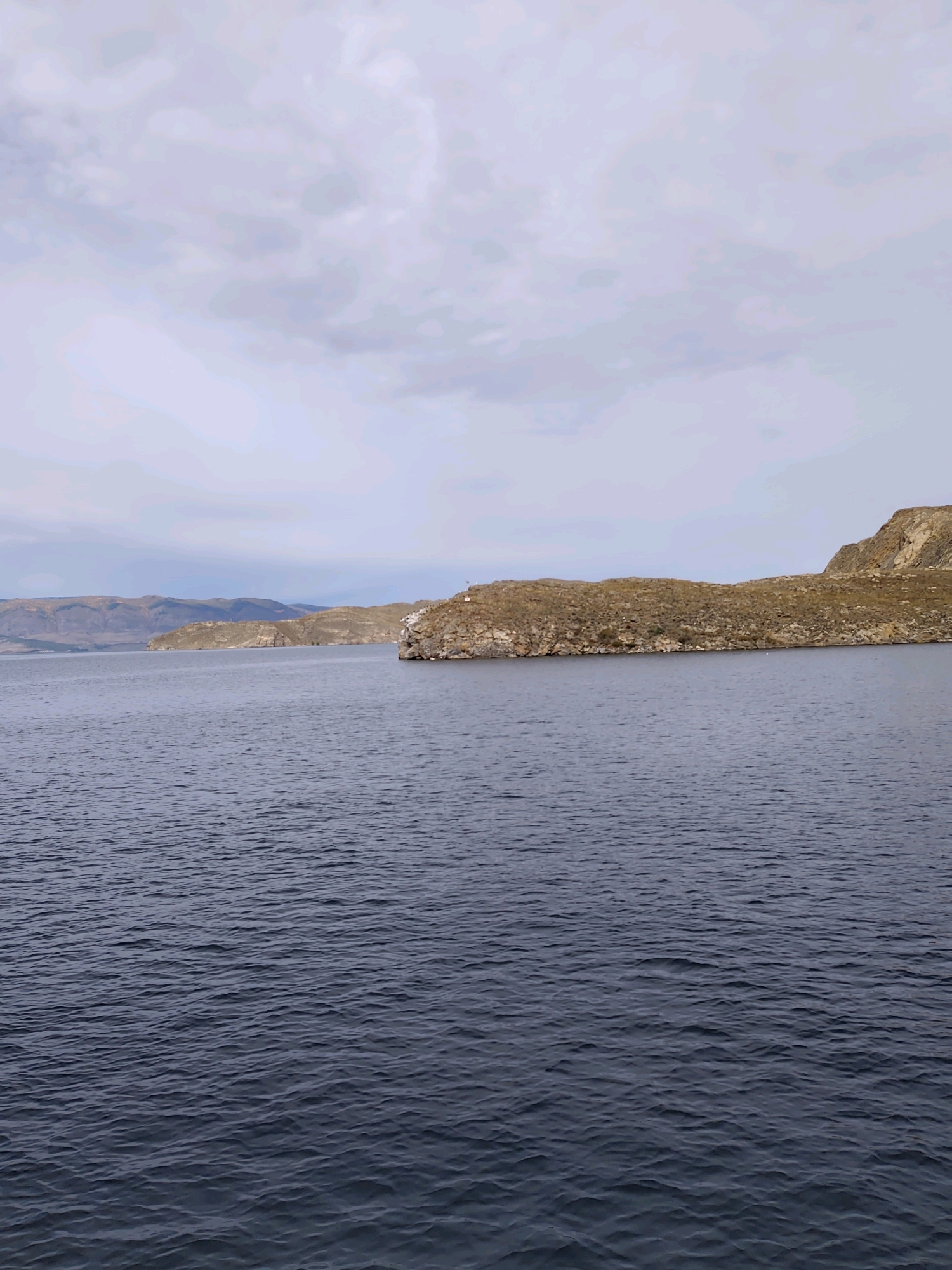 The image depicts a wide, deep blue body of water, likely a lake, with gentle ripples on its surface. In the background, a rugged, dry, and mountainous landscape stretches across the horizon. The terrain is characterized by brown and tan hills, sparsely covered with low-lying vegetation. The sky above is overcast with a layer of pale, cloudy formations. In the mid-ground, a rocky peninsula juts out into the water, topped with more of the arid, brown vegetation. A small, white lighthouse-like structure is visible on the very edge of this peninsula. The overall scene conveys a sense of vastness and natural, untamed wilderness. It is not possible to determine the specific location within the city of Khuzhir, Russia, based on the provided image alone.