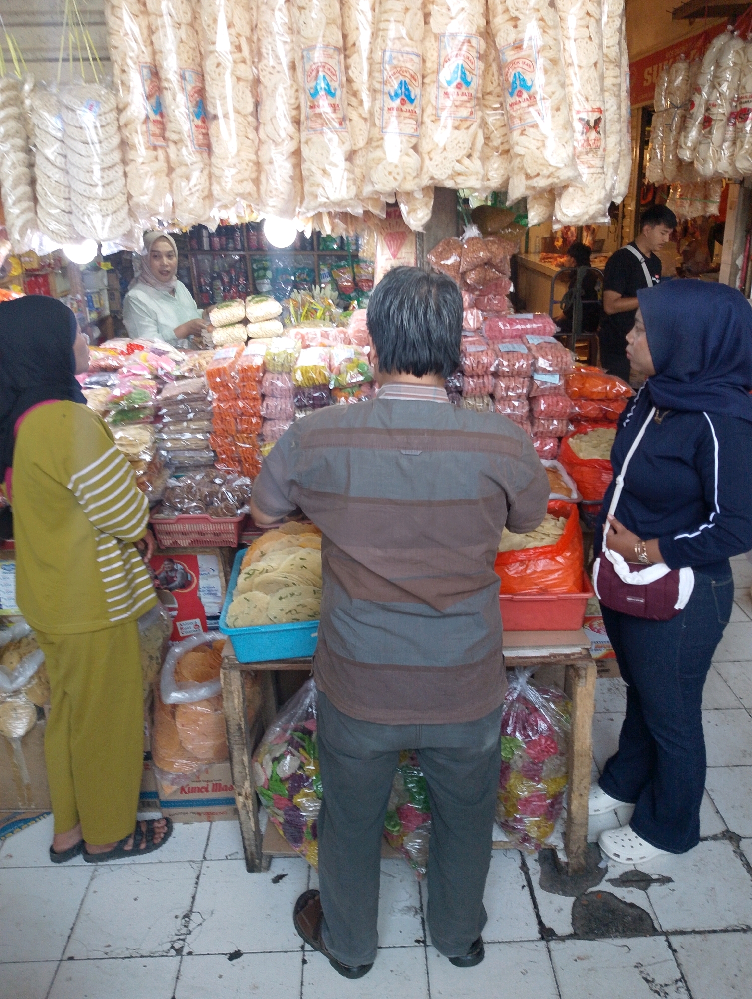 In Lembang, Indonesia, a street vendor displays an array of bagged snacks and crackers. The scene is bustling with activity, with customers interacting with the vendor. A woman in a yellow hijab and a bright yellow outfit, possibly a customer, is seen from the back, looking at the goods. A vendor, also a woman wearing a light green hijab and a light green top, is behind the counter, attending to a customer. Another customer, a woman with a dark blue hijab and a dark blue jacket, is in the foreground, facing the products. A man in a grey and brown striped shirt and dark pants is standing in the middle, looking towards the vendor. He appears to be a customer or perhaps an assistant. The market stall is filled with various food items, neatly arranged on shelves and in baskets. Bags of crackers hang from the ceiling, adding to the vibrant atmosphere. The floor is tiled with a grey and white pattern, showing some wear and tear, indicative of a busy public space. The overall impression is a typical Indonesian market scene, full of life and local flavor.