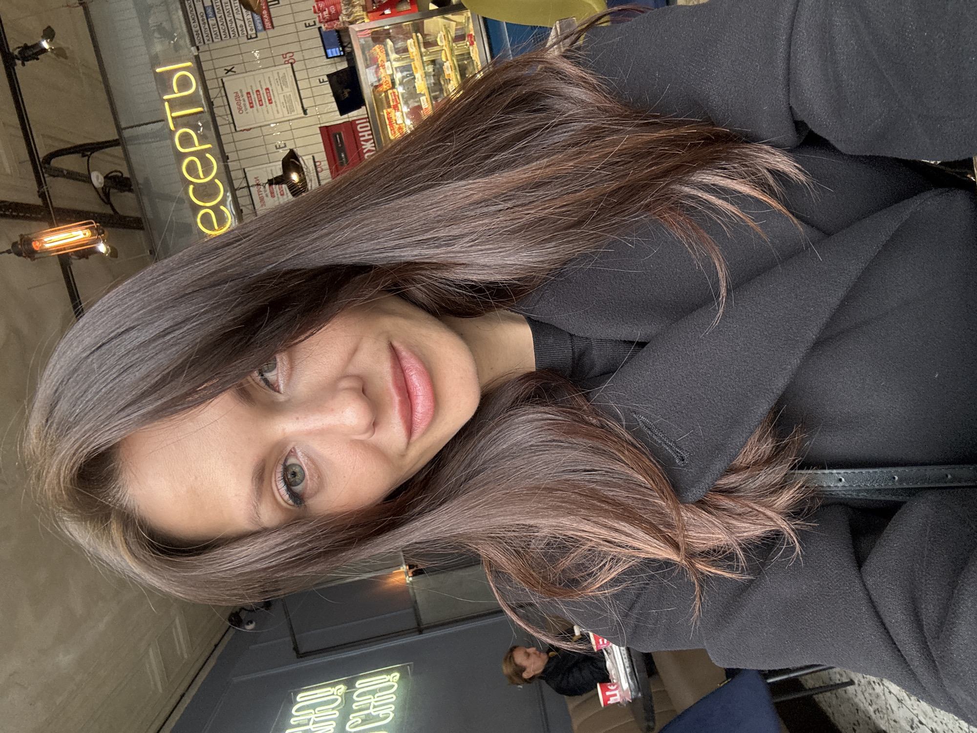 A woman is taking a selfie indoors, likely at a cafe in Saint Petersburg, Russia. She has long brown hair and is wearing a black coat. In the background, there's a neon sign in Russian reading "десерты" (deserty), meaning "desserts," a pastry display case, and other cafe-related signage. There's a glimpse of another person seated in the background.