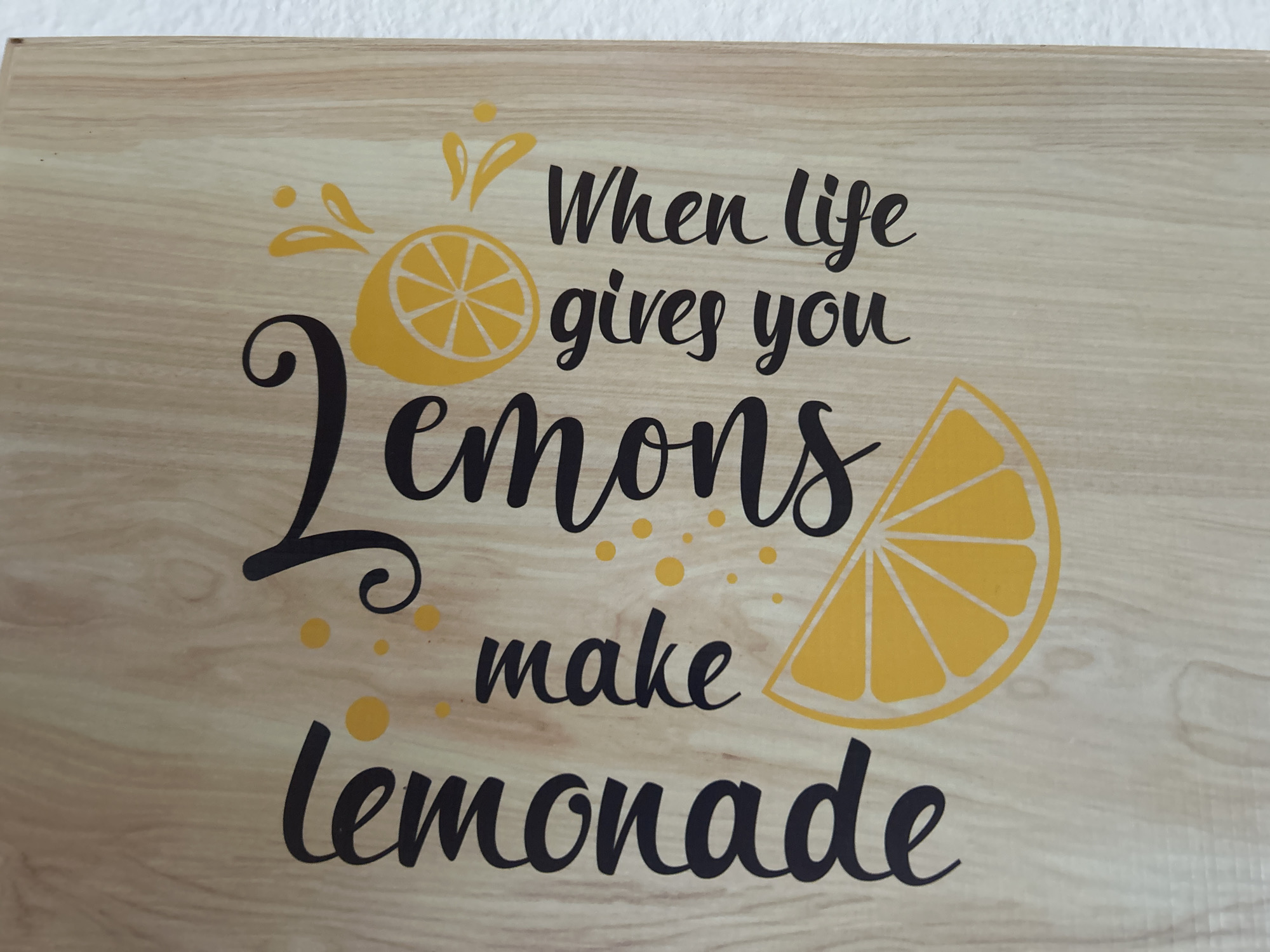 The image displays a close-up of a wooden surface with a quote and lemon illustrations. The quote reads "When life gives you lemons make lemonade" in a stylized font. A large "lemons" is prominently featured, with a lemon slice on the right and a whole lemon with splashes on the left. The overall aesthetic is cheerful and whimsical. This item appears to be a decorative piece, possibly a sign or a part of a kitchen accessory. There are no people or animals present. The environment is a neutral, indoor setting, with a white wall visible at the top. The object is situated in Detmold, Germany.