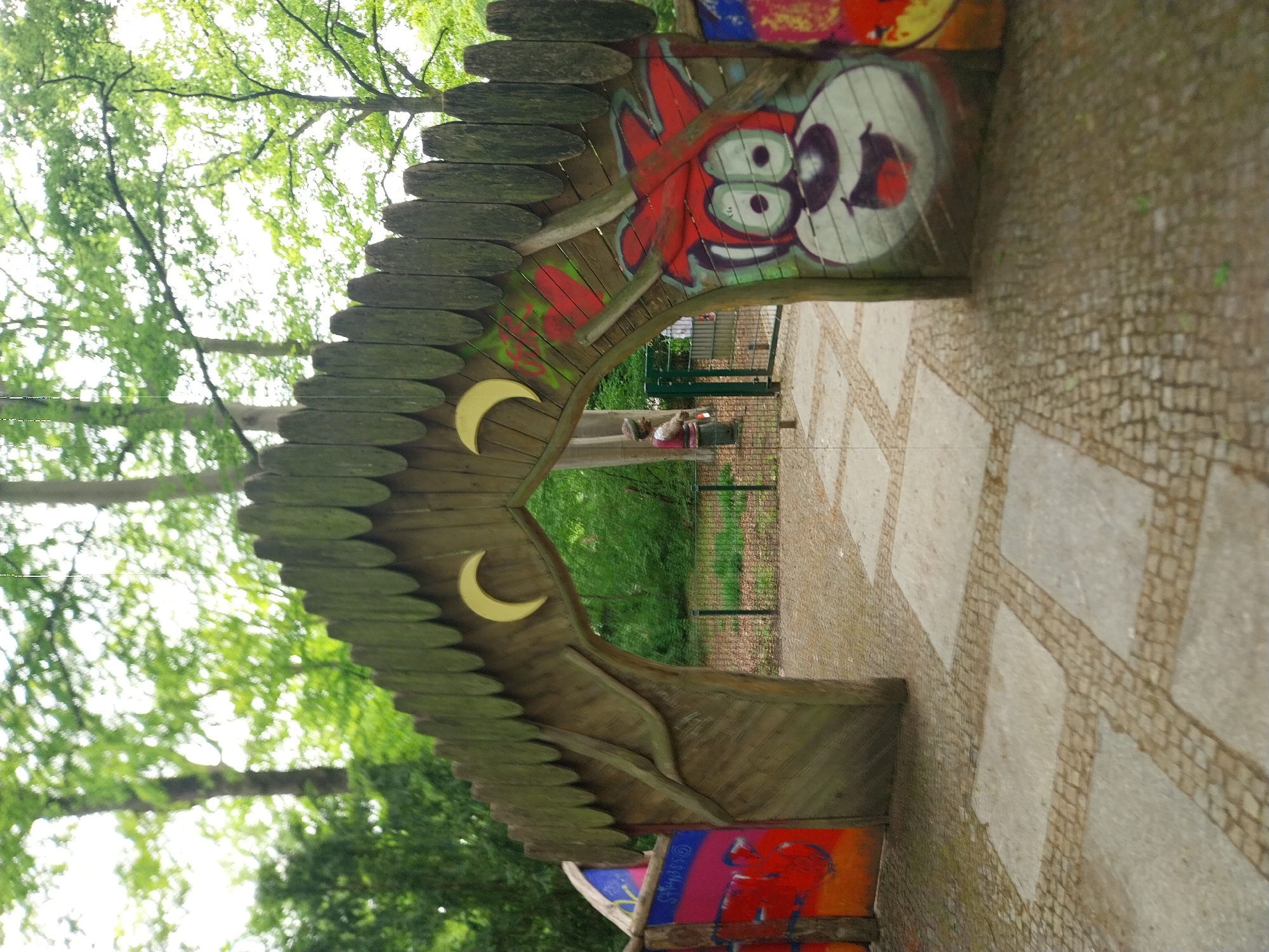 The image shows a wooden archway in a park, potentially in Berlin, Germany. The archway is decorated with two crescent moons and graffiti-style artwork featuring cartoonish faces and colorful designs. The path leading through the archway is paved with a combination of square stones and smaller cobblestones. A wire fence is visible beyond the archway, with trees and green vegetation in the background. A person is sitting on a bench beyond the archway.