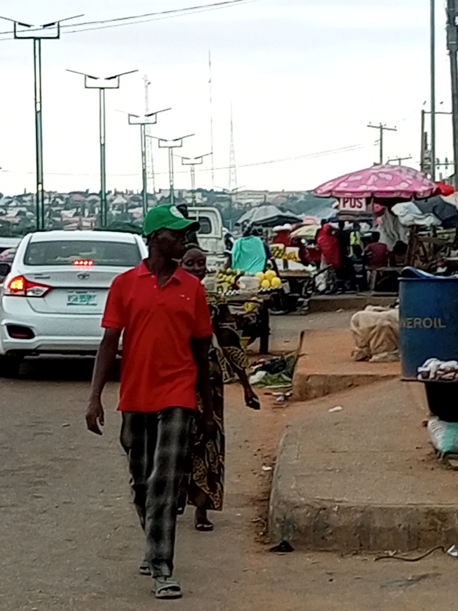 This image depicts a street scene in Gombe, Nigeria, likely near a market. In the foreground, a man wearing a red polo shirt and a green cap walks towards the viewer, followed by a woman in a patterned dress. Behind them, a white car is parked. The background reveals a bustling market area with stalls, umbrellas, and various goods. Structures and what appear to be streetlights line the horizon. The overall impression is of an active urban environment.