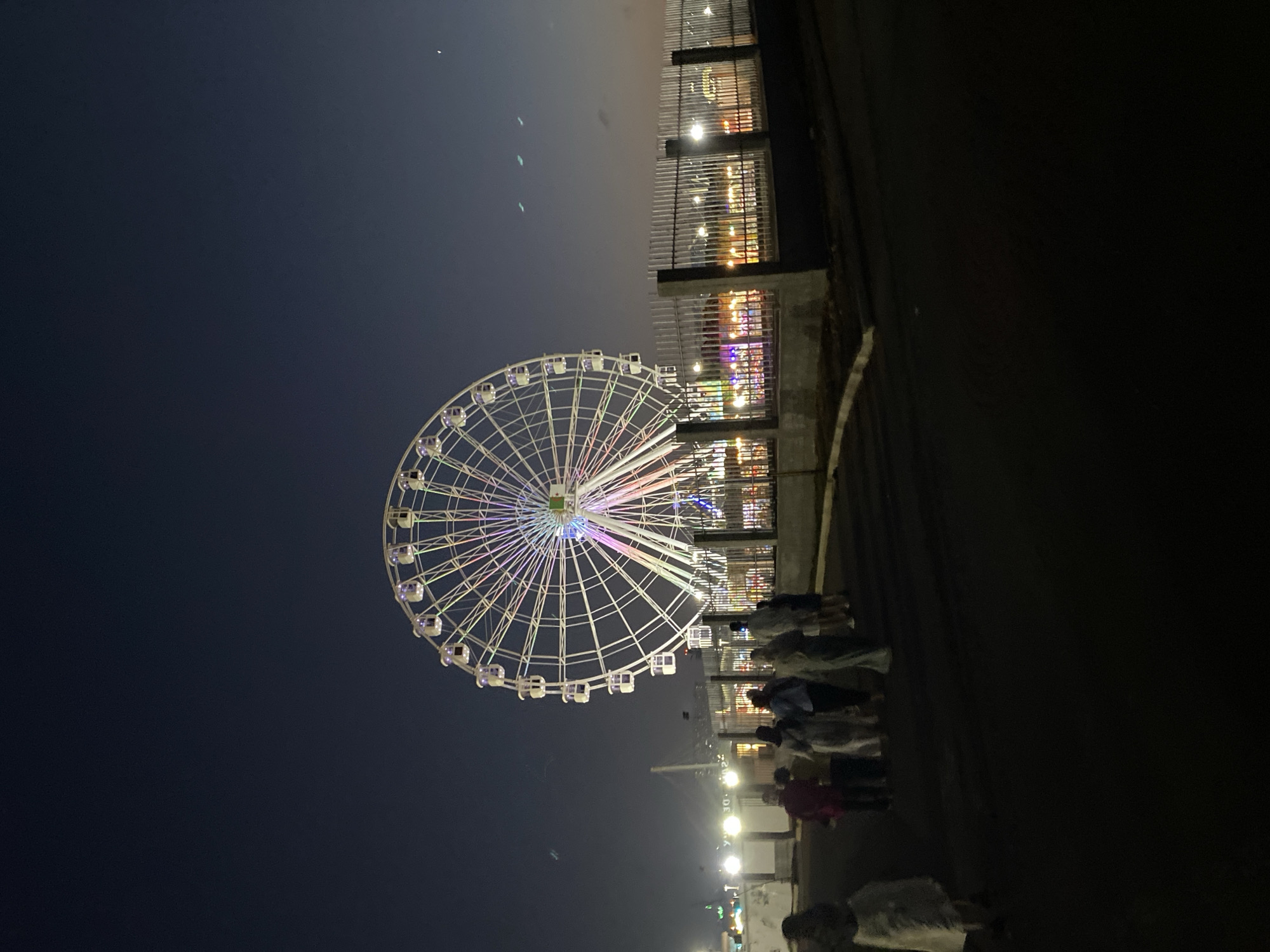 The image captures a Ferris wheel illuminated with colorful lights at dusk or night, situated in Hussein Dey, Algeria. A group of people, dressed in light-colored clothing, are walking on a paved path in the foreground, with their backs to the camera, towards the amusement area. The Ferris wheel is the dominant structure, a large white circular frame with numerous cabins, positioned behind a metal fence. In the background, city lights are visible, along with what appears to be a bridge or large structure. The sky is a deep blue, transitioning into a darker hue, indicating the time of day. The overall scene suggests an outdoor entertainment venue with a festive atmosphere.