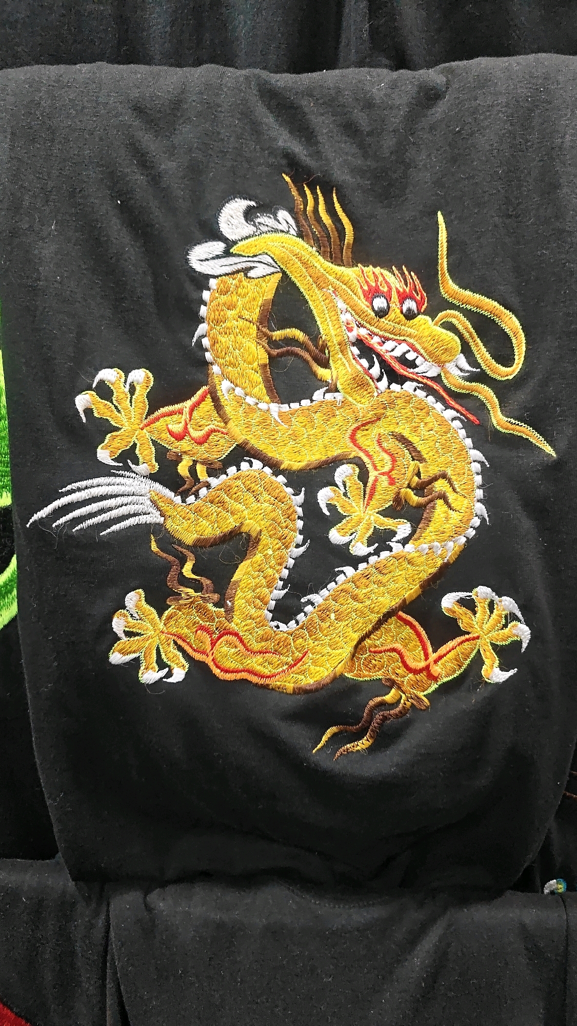 This is a close-up, eye-level, medium shot of a gold embroidered dragon on a black t-shirt. The dragon is coiled and facing the right side of the frame. It has yellow scales, red eyes, and sharp white claws. Red and white embroidery outlines the dragon's body. The fabric of the t-shirt is wrinkled and textured. The background is dark and blurred. The image does not provide any information about the city of Anjuna, India.