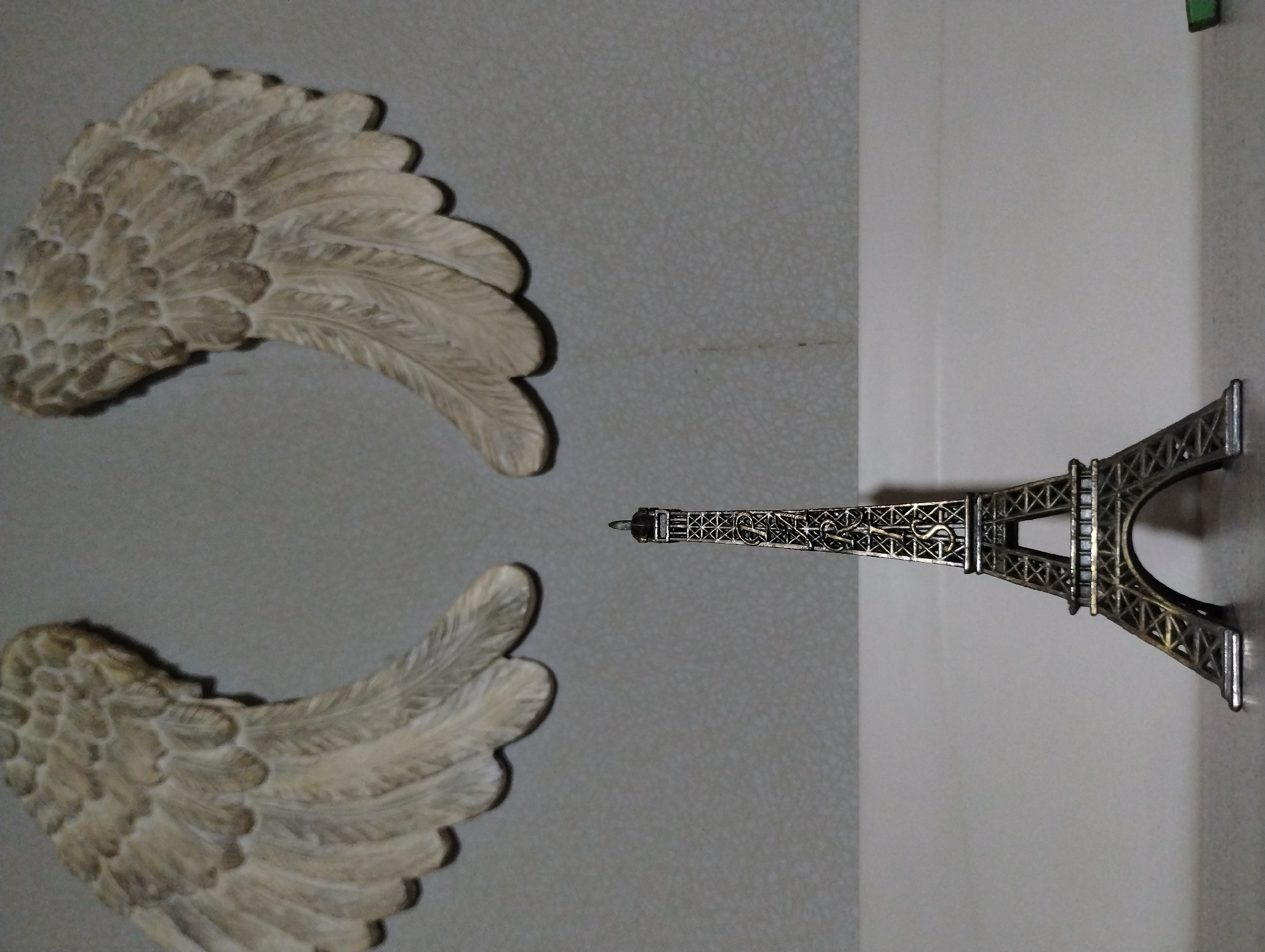 The image presents a static composition of decorative objects, viewed from a slightly elevated perspective. Two ornate, wing-shaped figures, resembling angel wings, dominate the left side of the frame. They are an off-white or light beige color with visible texture detailing individual feathers, and appear to be crafted from a solid material like plaster or resin. One wing is positioned in the upper left, and the other in the lower left, creating a symmetrical arrangement. Both wings cast distinct shadows on the underlying surface.
To the right of the wings, a miniature metallic replica of the Eiffel Tower stands vertically. The figurine is dark grey or bronze, featuring intricate structural details of the famous landmark's latticework. It is positioned upright on a lighter, plain off-white surface that contrasts with the textured grey surface beneath the wings. A faint seam or corner is visible where these two surfaces meet on the right side of the image.
The background comprises two distinct areas: a textured, light grey surface on the left, and a smoother, lighter grey or white surface on the right. The lighting suggests an indoor setting, with soft shadows indicating a light source from above or the side. There is no activity or event taking place, as the objects are static decorations. No people are visible, and there is no discernible text on any of the objects or the background. The scene is situated in Roza, Bulgaria.