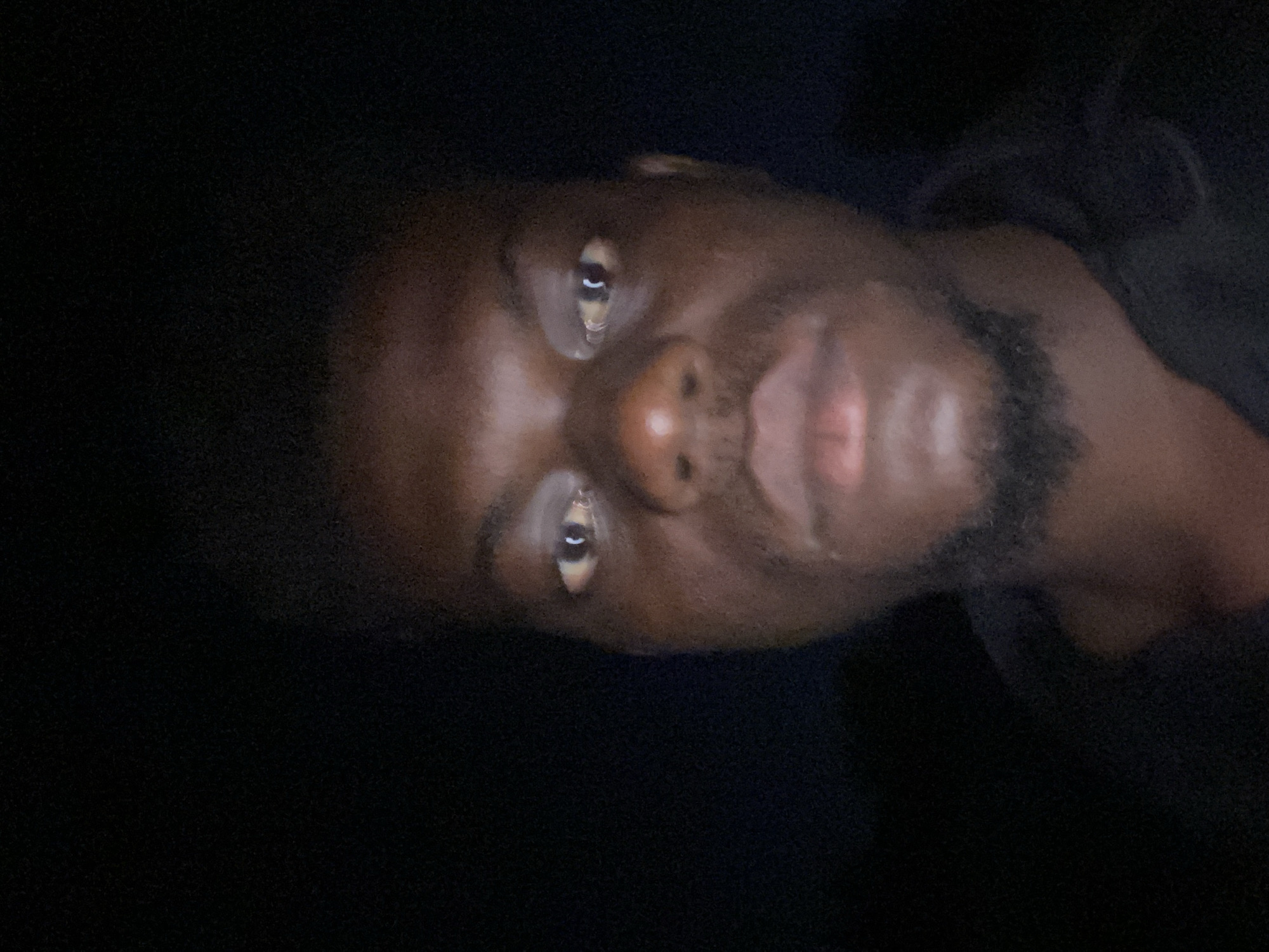 A close-up, poorly lit portrait of a dark-skinned man with a beard. The background is dark. Location: Unknown, Unknown.