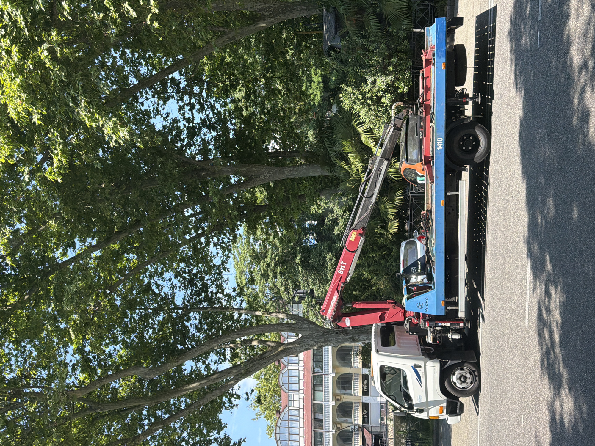 On a bright, sunny day, a white tow truck with a red crane is parked on the right side of a paved road. The truck has a blue flatbed. A silver car is partially visible on the flatbed, secured for towing. The truck is parked in front of a multi-story building with arched windows and a red roof. Lush green trees with large trunks and dense foliage line the background, suggesting a park or boulevard setting. The ground beneath the truck is shaded by dappled sunlight filtering through the trees. The overall scene is urban, likely within Sochi, Russia, given the presence of a building with architectural elements and signage that could be Cyrillic. The sky is clear and blue.