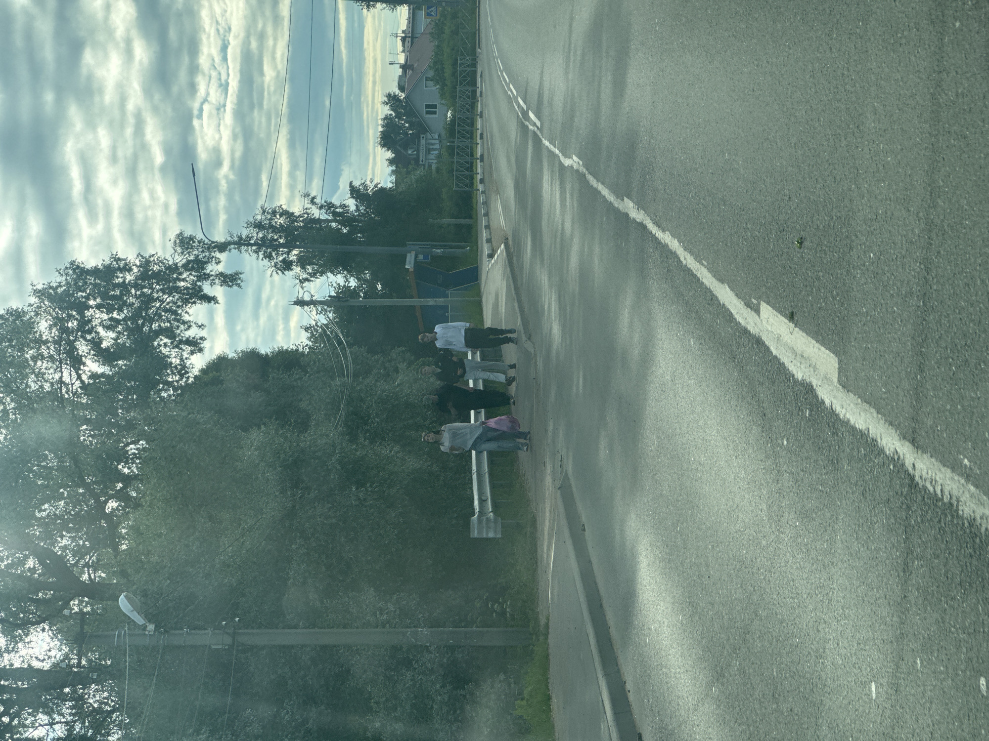 The image shows a street scene in Annino, Russia. The foreground features a paved road with a white line marking. The background is dominated by lush green trees and a cloudy sky. Several people are visible standing on the side of the road, some appearing to be waiting. One person is wearing a white shirt and dark pants, while others are dressed in darker clothing. One individual is carrying a pink bag. There are streetlights and power lines visible, suggesting an urban or suburban environment. The sunlight filtering through the clouds indicates it is daytime, likely late afternoon or early evening given the soft lighting and shadows. The overall atmosphere appears to be calm and ordinary, depicting everyday life on a road in a residential area. There are some buildings visible in the distance, nestled among the trees. No text is clearly legible in the image.