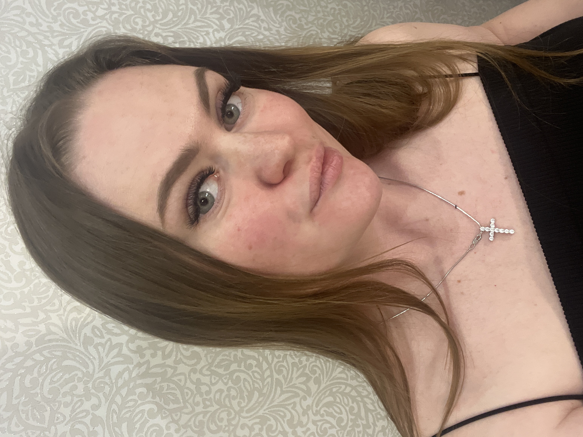 This is a close-up, tilted portrait of a young woman taken in an indoor setting. The subject is looking directly towards the camera with her head angled. She has long, medium brown hair, which frames her face and extends across her shoulders. Her eyes are light-colored, possibly green or hazel, and are emphasized by prominent, long eyelashes and well-defined eyebrows. Her fair skin shows some natural redness on her cheeks. Her lips are slightly parted in a subtle pout.
She is wearing a black top with thin spaghetti straps, visible on her shoulder and upper chest. A silver-toned necklace with a sparkling cross pendant is worn around her neck, resting on her décolletage.
The background features a light-colored surface with a subtle, ornate pattern of swirling white and grey motifs, resembling wallpaper or fabric. The lighting is soft and even, typical of an indoor environment. No specific time of day or weather is discernible. There is no visible text in the image. The location context provided is Sevastopol, Ukraine.