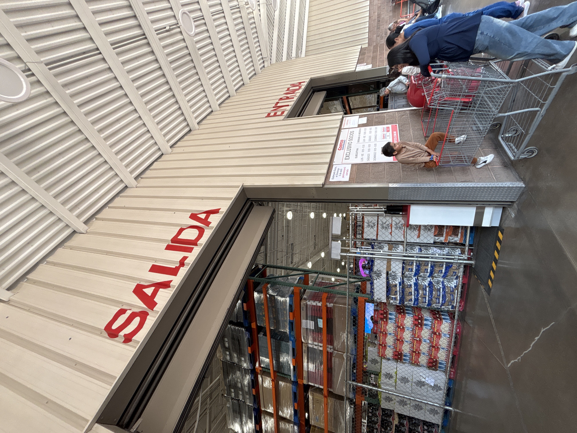 The image shows the entrance and exit of a Costco store in Puebla, Mexico. The entrance sign reads "ENTRADA" in Spanish, and the exit sign reads "SALIDA" in Spanish. People are seen at the entrance, some pushing shopping carts. There is a sign with the Costco logo and membership information (EXCLUSIVO SOCIOS), including hours of operation for both Executive and Gold Star/Business members. Inside the store, shelves are stacked with merchandise. The overall environment is that of a busy retail store entrance.