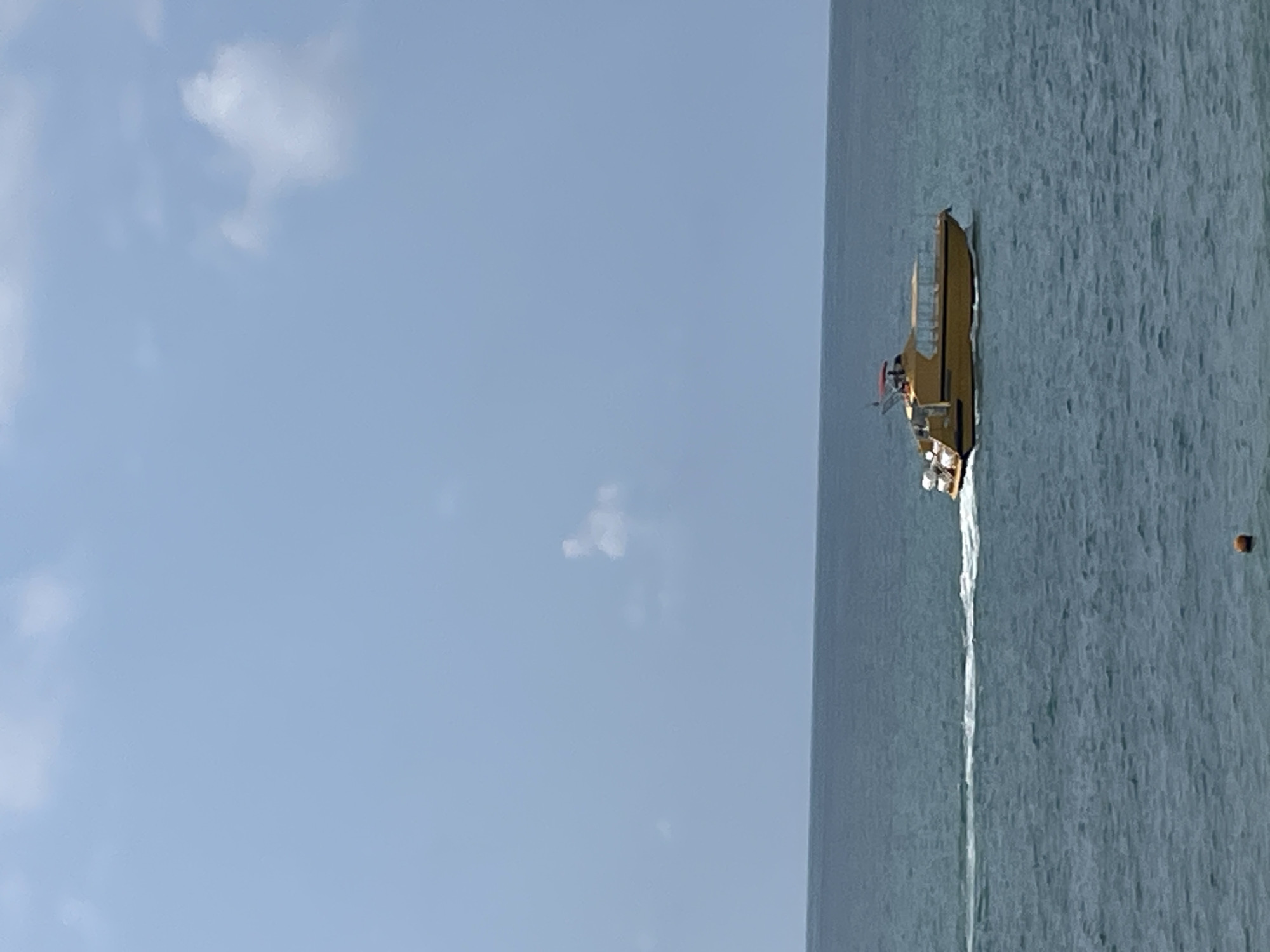 A yellow passenger boat is traveling across the calm, blue sea under a clear sky with scattered white clouds. The boat has a red roof and appears to be moving at a moderate speed, leaving a white wake behind it. There is a small orange buoy floating in the foreground. The image depicts a serene maritime scene, likely along the coast of Sochi, Russia.