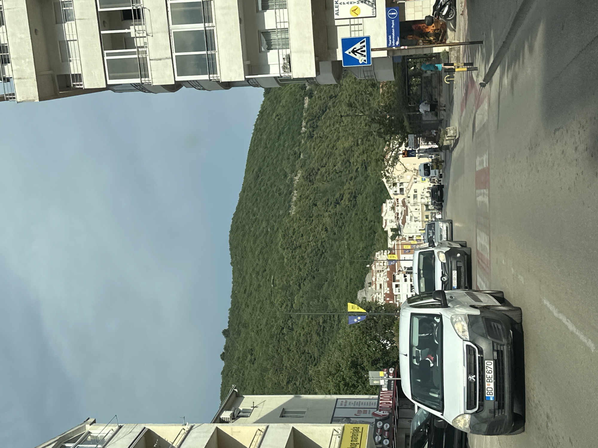 The image captures a street scene in Budva, Montenegro. In the foreground, a white Peugeot van with license plate BD BE 670 is prominently featured, driving down a paved road. Several other vehicles are visible on the road, including a white van behind the Peugeot and a silver car further down. The road has pedestrian crossings with red and white stripes.
To the left of the road, a modern, multi-story building with balconies and large windows dominates the view. A yellow sign with black text is attached to the side of this building. Further down the street, on the right side, another multi-story building with balconies and windows is visible. A blue sign with a white pedestrian symbol is mounted on a pole in front of this building, along with a sign indicating a "Tourist Information Point."
Behind the buildings, a densely forested green hill rises majestically. Scattered across the hillside are several white buildings with red roofs, typical of Mediterranean architecture. Two flags, one blue and yellow, and another yellow flag with a star, are visible on poles in the middle ground, suggesting a public space or gathering area.
The sky is overcast with a pale blue hue. The overall impression is a bustling urban environment nestled against a natural, green backdrop. The time of day appears to be daytime, with ample natural light.
