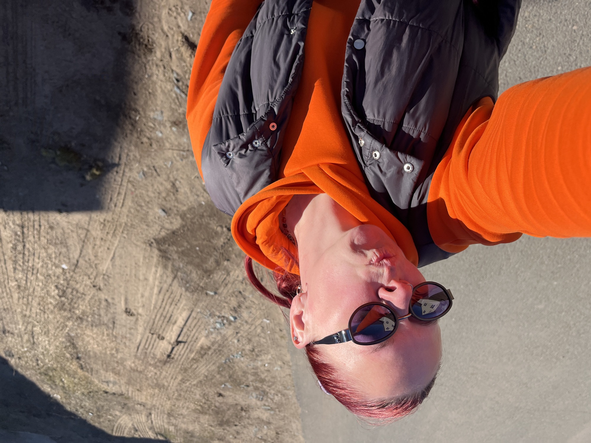 A person is shown from a high angle, seemingly in a selfie. They have light skin, red hair, and are wearing round sunglasses. They are dressed in an orange hooded sweatshirt and a dark grey puffer vest. The ground is partially visible, with a mix of rough dirt and smooth concrete or asphalt. Based on the reflection in the sunglasses and the type of ground seen, this could be a park or similar outdoor location. There is no explicit information about the city of Chaikovsky, Russia in the visual content.