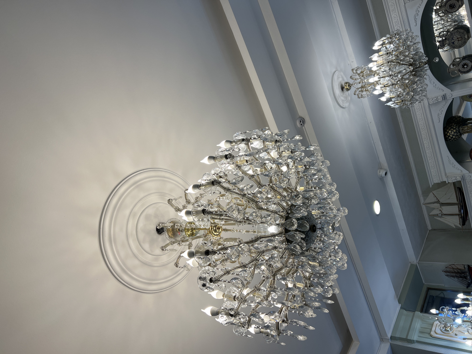 The image shows an interior view, likely taken from a low vantage point, featuring a ceiling with decorative elements. Two large crystal chandeliers dominate the scene. The chandeliers are complex, globe-shaped, with multiple light sources and crystal ornamentation. The ceiling is white with recessed lighting. Ornate molding designs surround the base of the chandeliers. The image was taken in Tbilisi, Georgia.