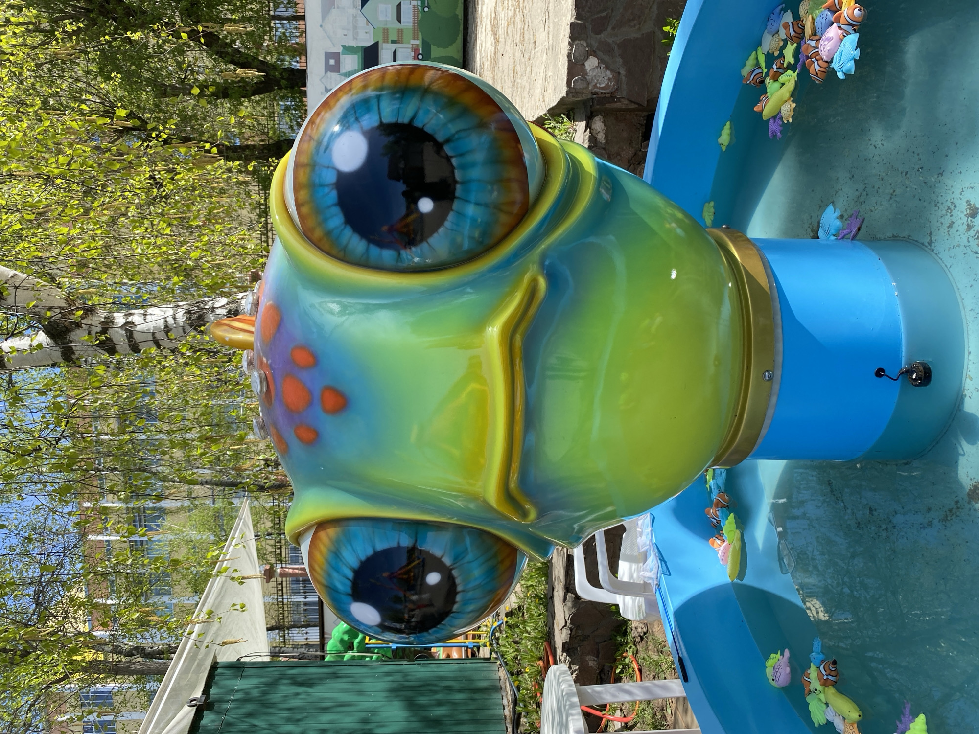 The image shows a close-up of a colorful, cartoonish frog head sculpture. The sculpture is primarily green and blue, with large, round eyes. The eyes have black pupils and blue irises with brown edges. The head has orange spots on the upper portion. The sculpture is part of a water feature, with a blue pool or basin surrounding it. Several toy fish are floating in the water. The background includes trees with green leaves, suggesting a park or outdoor setting. In the distance, buildings and other structures can be seen. The image was taken in the city of Izhevsk, Russia.
