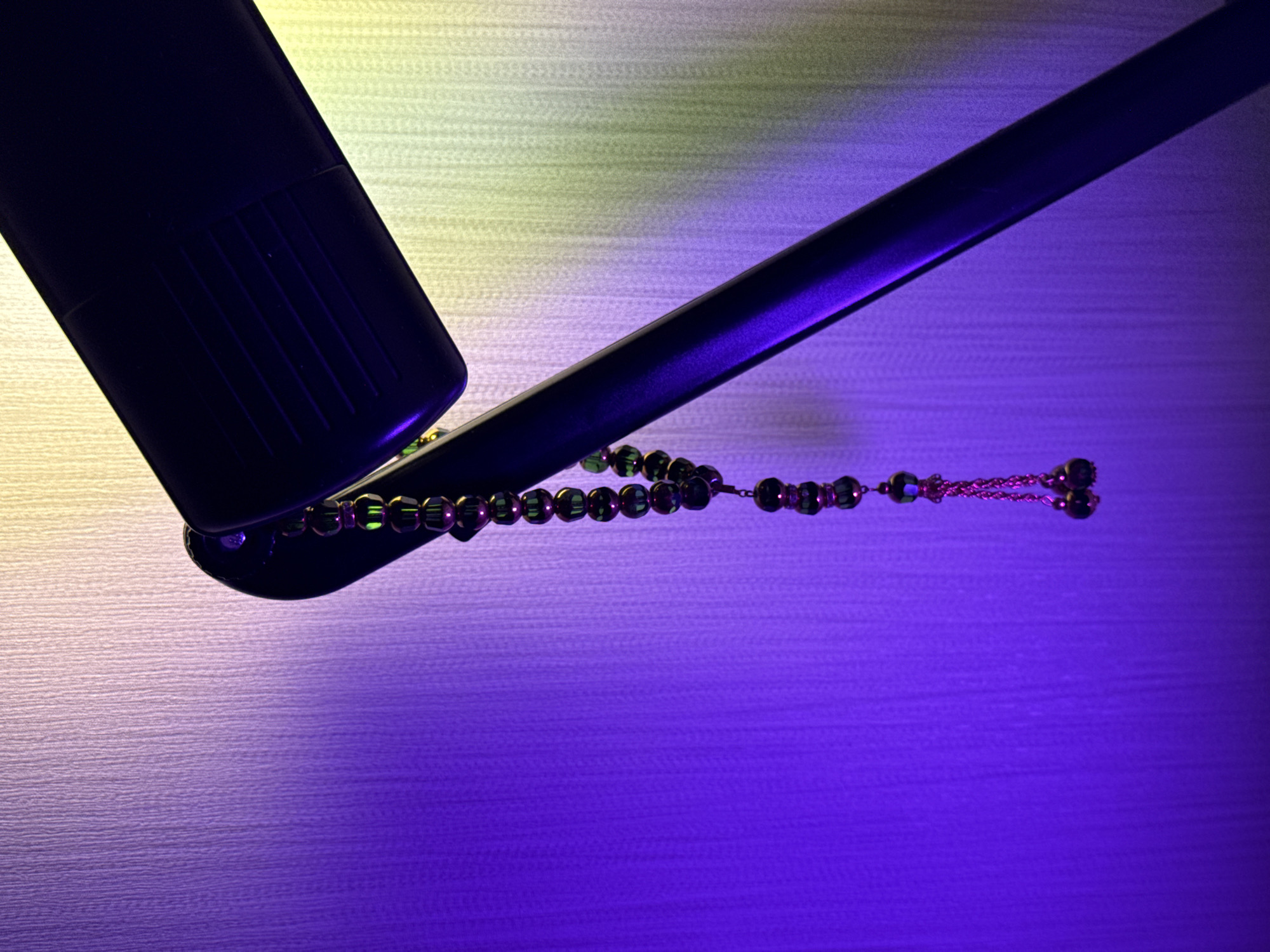 A close-up, low-angle shot shows a string of dark green prayer beads with gold accents hanging from the arm of a black desk lamp. The lamp is positioned in the upper right of the frame, with its head angled downwards. The background is a textured wall illuminated by purple light on the left and yellow light on the right, creating a gradient effect. The beads are the primary subject, with their chain and charms visible in detail. The scene is devoid of any people or overt human activity, suggesting an indoor, static setting. The lighting, color palette, and the presence of the prayer beads evoke a sense of quietude or contemplation.