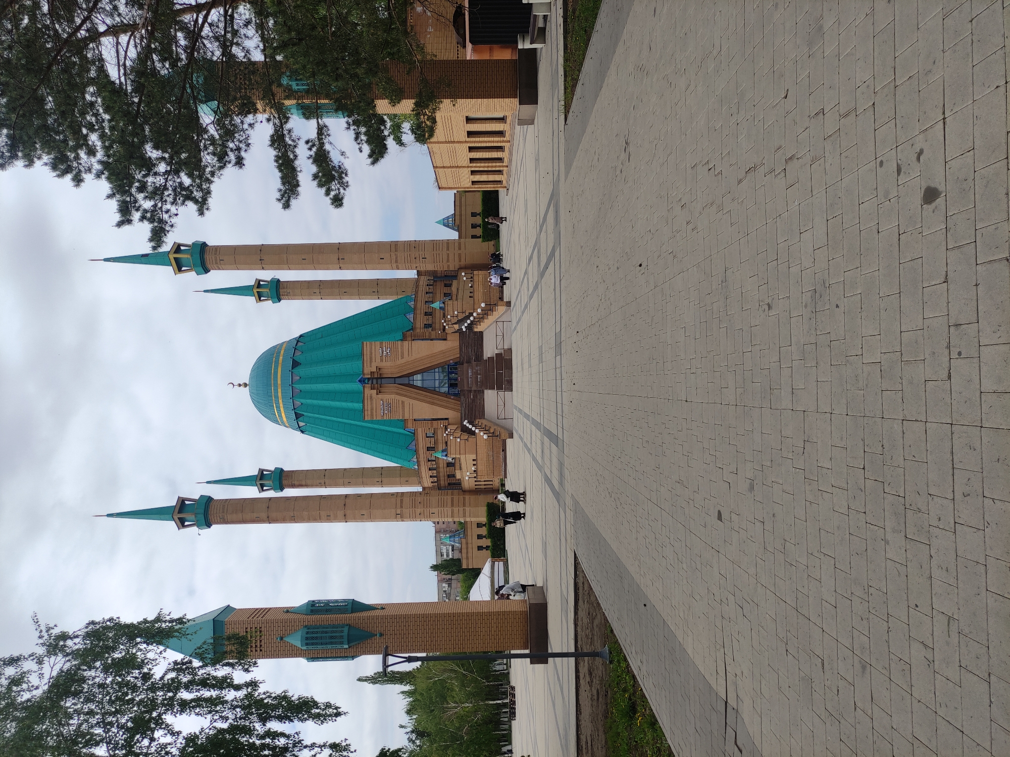 The image shows the Qolsharif Mosque in Kazan, Russia, not Pavlodar, Kazakhstan. The mosque features a large turquoise dome and four tall minarets, also with turquoise tops. The building is constructed primarily of light brown brick. A wide paved walkway leads towards the mosque entrance. Several people are walking on the walkway near the mosque. There are trees visible in the foreground and background, with an overcast sky above.