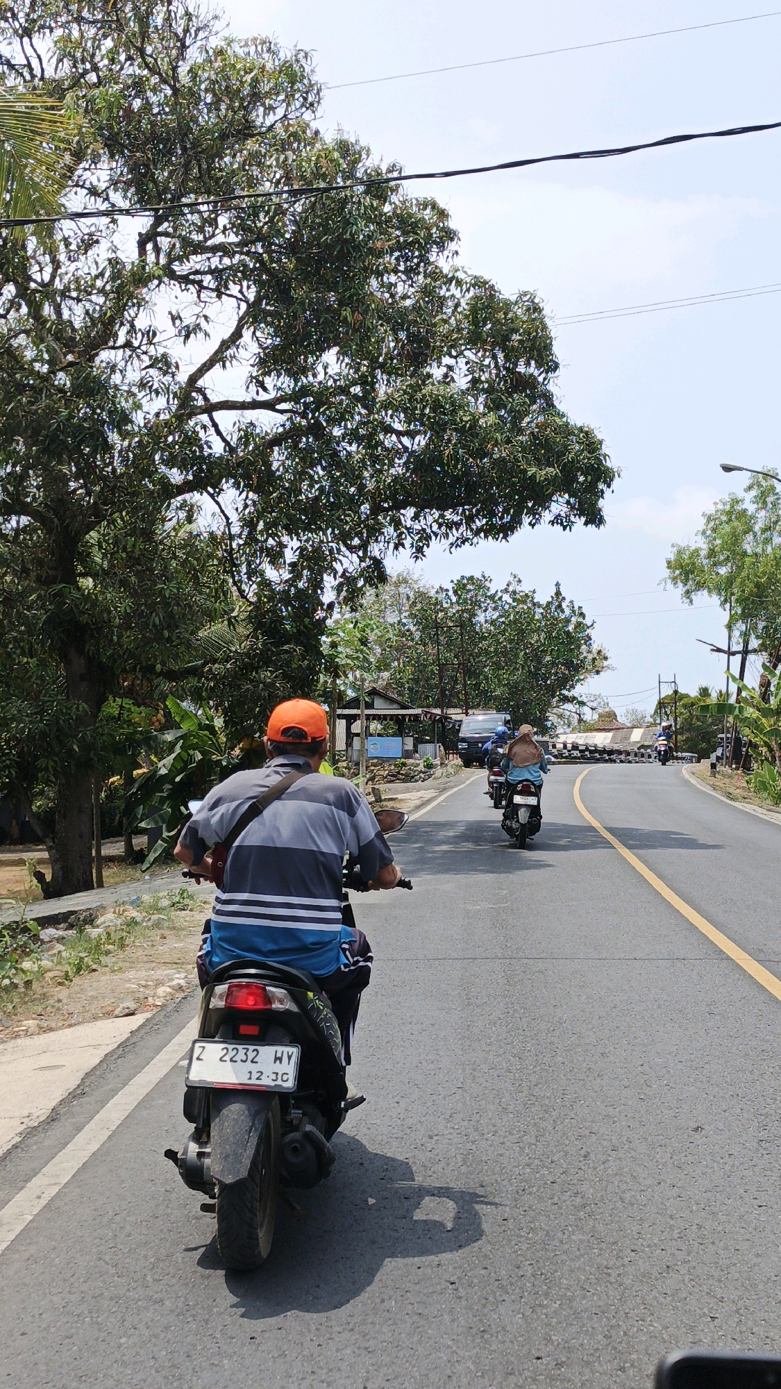 A person wearing an orange cap and a striped t-shirt is riding a motorcycle on a paved road. The motorcycle has a license plate with the number "Z 2232 WY" and "12.30". The rider is looking ahead, and the motorcycle is moving forward. In the background, other motorcycles and a van are visible on the road, as well as trees and buildings. The sky is clear and blue. The scene appears to be in a rural or semi-rural area of Cikembulan, Indonesia.