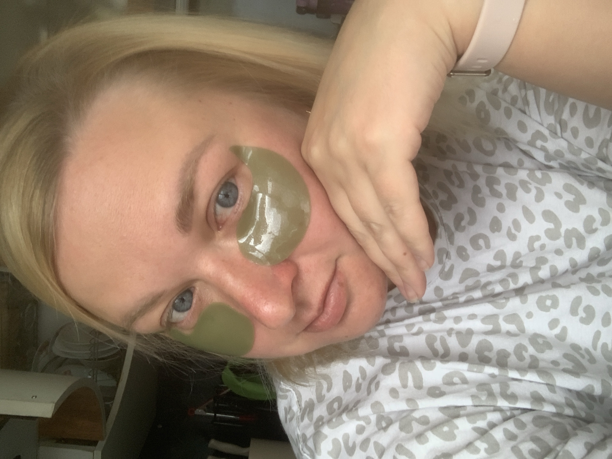 A fair-skinned woman with blonde hair and blue eyes poses for a selfie. She has two green under-eye patches on her face. She is wearing a white shirt with gray leopard print. A light pink smartwatch is on her left wrist. In the background are miscellaneous objects suggesting an indoor setting, perhaps a home. There is no verifiable information to confirm the location is Chaikovsky, Russia.