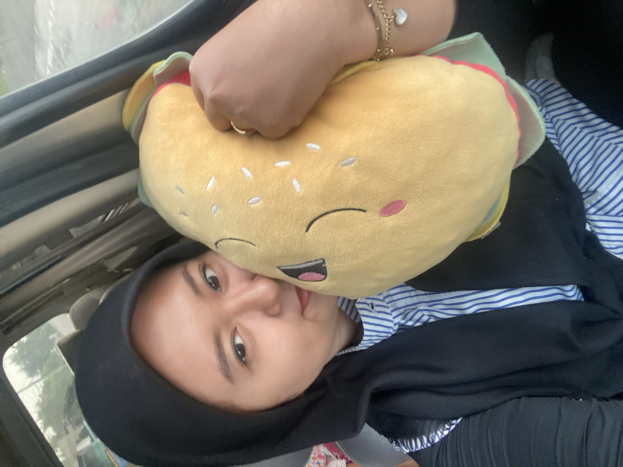 A young woman is holding a plush toy of a burger. She is wearing a black hoodie and a striped shirt. The location is inside a vehicle, and it is presumably in the city of Tlasih, Indonesia, given that this is the provided location. The woman is looking at the camera. A hand with a bracelet is visible at the top of the image.
