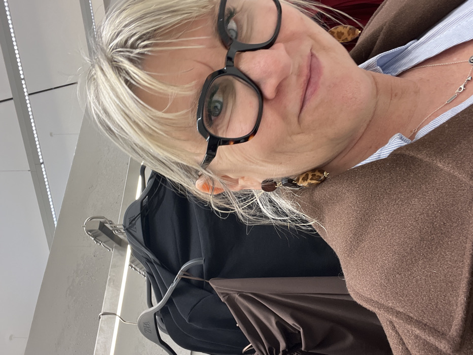 A close-up, selfie-style photograph depicts an adult female positioned within a ZARA retail clothing store in Saint-Jean-Bonnefonds, France. The female subject, appearing to be light-skinned with blond hair showing some graying and styled with bangs, occupies the foreground. She wears black, thick-rimmed, rectangular glasses with tortoiseshell details and has light blue eyes. Her facial expression suggests a faint smile or smirk. She is dressed in a brown or camel-colored coat over a light blue collared shirt. A silver-colored necklace with multiple small pendants, including a possible cloverleaf, is visible. She wears multi-component drop earrings, each consisting of a dark oval upper piece and a larger, elongated, leopard-print lower piece. The background reveals rows of clothing racks displaying numerous black garments and one brown garment, all hanging on gray plastic hangers branded with the "ZARA" logo. The interior environment includes a light-colored wall and a ceiling equipped with linear, bright LED light fixtures.