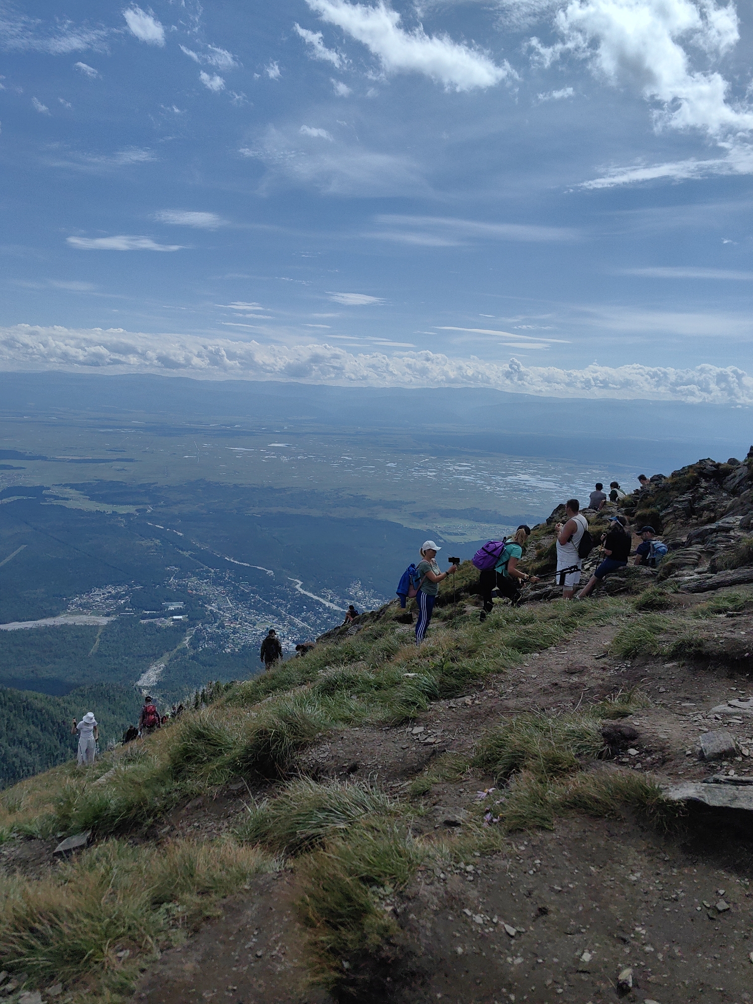This image depicts a group of hikers on a steep, rocky mountainside overlooking a valley with a town and agricultural fields. The sky is bright blue with scattered white clouds. The foreground shows a well-worn dirt path with sparse green grass and rocks. Several people are visible on the path and rocky slopes, some standing, some sitting, and some appearing to be taking pictures or resting. They are dressed in casual hiking attire, with backpacks.

In the middle ground, a town with numerous buildings is spread across the valley floor, bisected by a winding river. Beyond the town, the landscape transitions into a vast expanse of green fields, some of which appear to be flooded or contain bodies of water. Mountains form the distant backdrop, softened by haze. The overall impression is of a scenic vista from a high vantage point, likely a popular hiking trail in the vicinity of Arshan, Russia.