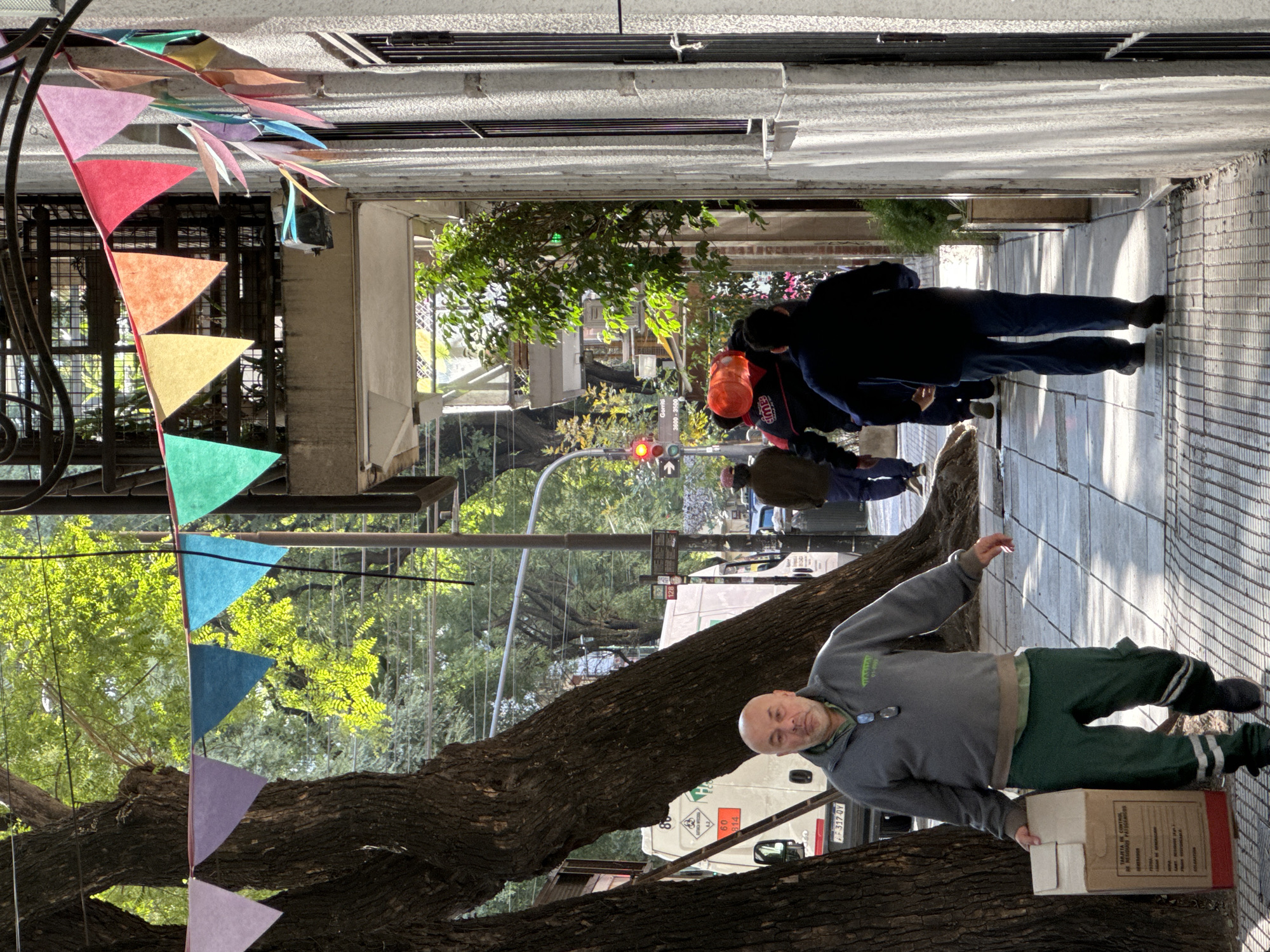 The image depicts a street scene in Buenos Aires, Argentina. The view is oriented along a narrow sidewalk lined with buildings on either side. A large tree trunk extends across the lower half of the frame, with triangular bunting suspended above. A man in a gray sweatshirt and green pants is near the tree, holding a cardboard box. Further down the sidewalk, a group of people are walking. A street with a traffic light is visible at the end of the sidewalk. A truck is parked behind the tree.