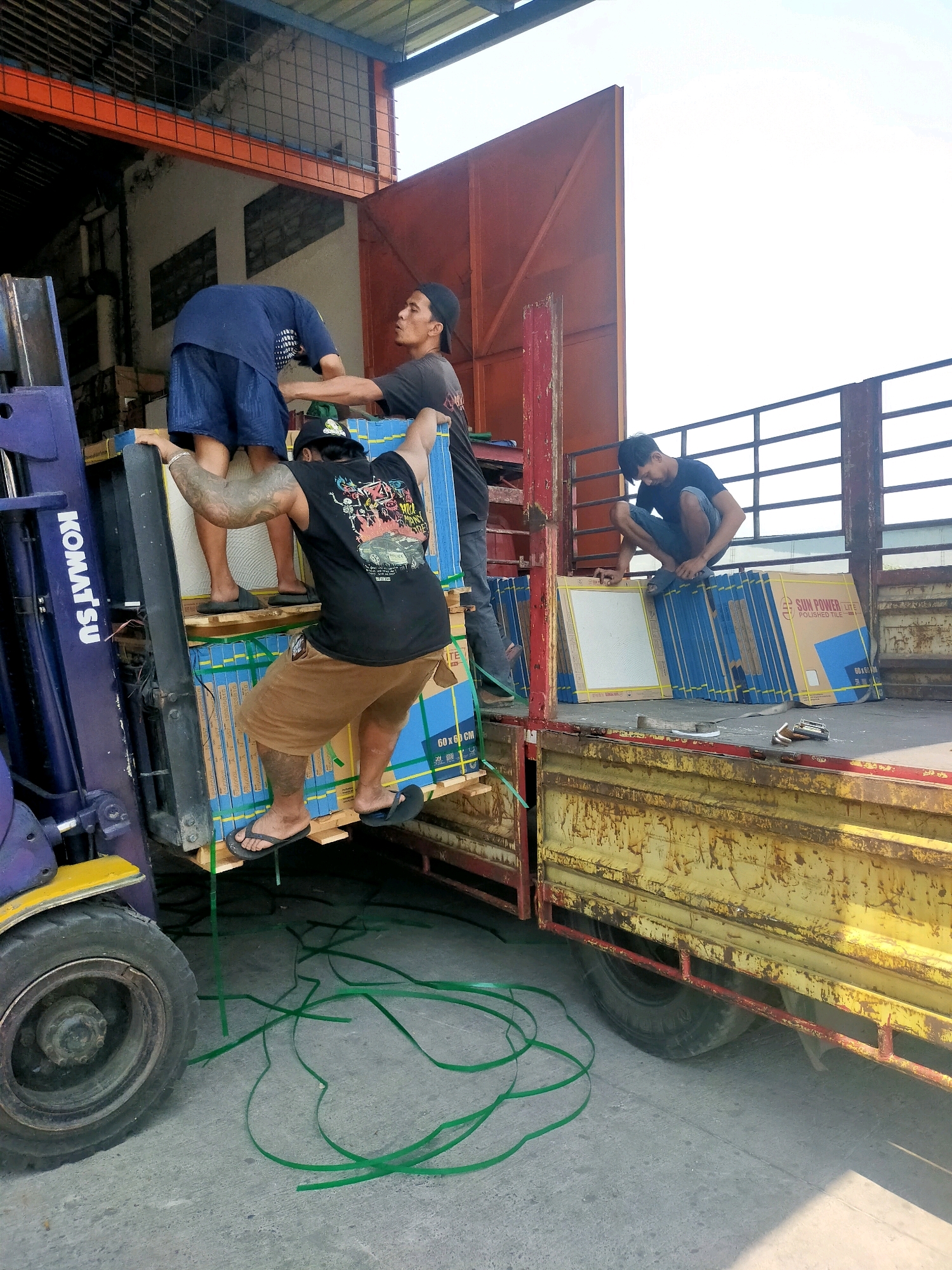 In Mlaten, Indonesia, at a loading area, three men are seen actively engaged in unloading stacked boxes of tiles from a forklift onto the flatbed of a yellow, weathered truck. The boxes are predominantly blue and yellow, with branding indicating "SUN POWER POLISHED TILE." One man, wearing a black t-shirt and brown shorts with tattoos on his arm, is climbing onto the pallet of tiles while steadied by a forklift operator wearing a blue shirt and shorts. Another man in a black t-shirt and dark pants is crouched on the truck, organizing the tiles. A third man in a dark t-shirt and blue jeans is also on the truck, observing the process. Green plastic strapping material is scattered on the ground around the forklift and truck. The environment appears to be an outdoor industrial or storage area, with a partially covered structure overhead and a bright sky visible in the background.