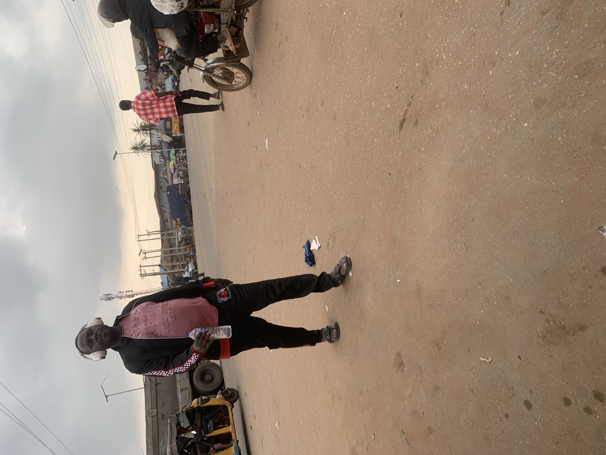 A man wearing a pink t-shirt, a black jacket with checkered sleeves, and black track pants stands facing forward in the foreground, holding a bottle of water in his right hand. He is also wearing white headphones and dark sandals. To his left, a yellow tricycle is parked. In the background, a large truck is visible, as well as a motorcycle with a rider wearing a helmet. Several buildings and streetlights line the background, suggesting an urban environment. The ground is a dirt or sandy surface, common in many Nigerian cities, and the sky is overcast with a slightly hazy appearance. The scene appears to be a busy street or market area in Shagamu, Nigeria, likely during the daytime.
