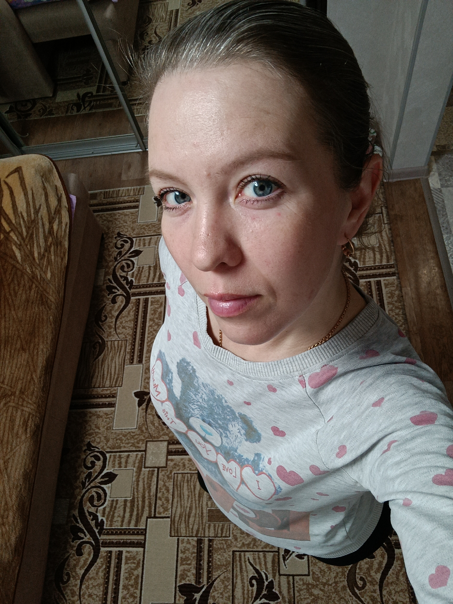 The image shows a woman with fair skin, light blue eyes, and blonde hair pulled back. She is wearing a light grey long-sleeved shirt with pink heart patterns and a design on the front. A gold chain necklace is visible around her neck, and she is wearing hoop earrings. She is looking directly at the camera with a neutral expression.
The background includes a patterned carpet in shades of brown and tan, a brown couch with a patterned blanket on the left side, and part of a sliding door closet in the upper part of the image. The photo appears to be taken from a high angle, looking down on the woman.
The location is Chaikovsky, Russia, based on external metadata.