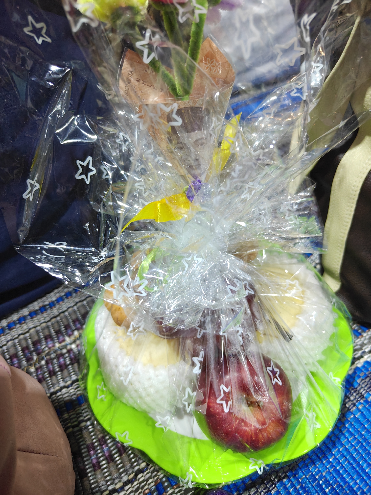 An overhead shot shows a gift basket wrapped in clear cellophane with white stars. Inside the basket, a bright green plate holds white rice balls, a red apple, and grapes. The basket is sitting on a patterned blue and brown mat. In the background, a dark blue cloth with white stars is visible. The gift basket is a custom-made arrangement, likely a bento box or a gift for a special occasion, a common sight in Cermee, Indonesia, where such personalized gifts are popular.