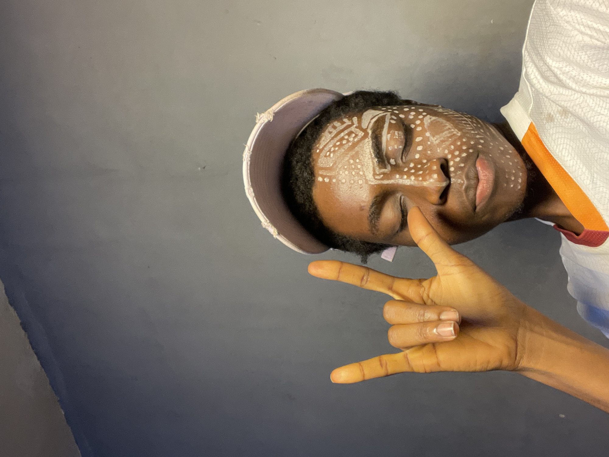 A young, dark-skinned person with closed eyes is photographed from a low angle against a gray background. They are wearing a light-colored baseball cap and a white and orange top. The person's face is decorated with white paint designs. They are making the "rock on" hand gesture, with their index finger and pinky extended. The location is Nkpor, Nigeria.