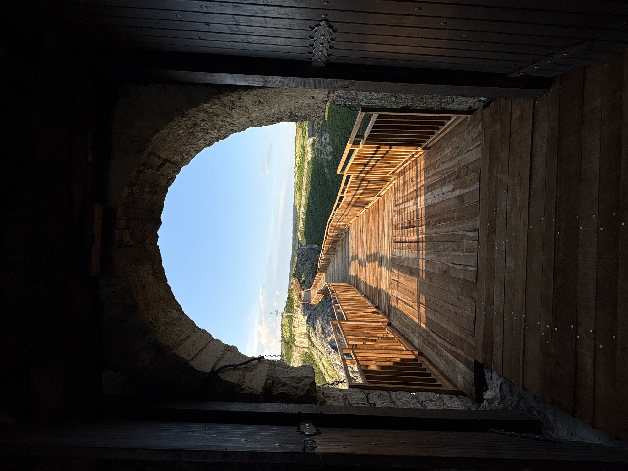 The image captures a detailed view through a dark, stone archway, opening onto a brightly lit, curving wooden walkway in the vicinity of Provadia, Bulgaria. The foreground is dominated by the shadowed interior of an architectural structure, framed by a rough-hewn stone arch overhead and partially open dark wooden panels on the left and right, featuring decorative metal hardware on the right panel. The interior floor consists of dark wooden planks.
Beyond the archway, a long, wide wooden walkway extends into the distance, constructed from light brown planks. Wooden railings with vertical balusters line both sides of the walkway, which gently curves to the right as it recedes. Strong, directional sunlight illuminates the walkway and railings from the right, casting long, parallel shadows of the balusters across the wooden surface, indicative of a low sun angle.
The walkway is situated along a natural landscape featuring steep, verdant cliffs and hillsides covered in green foliage, overlooking a deep valley also densely covered with greenery. The sky above is a clear blue with sparse, white cumulus clouds. In the far distance, prominently positioned on an elevated landform, a large white cross structure stands against the sky. No individuals are present in the captured scene.