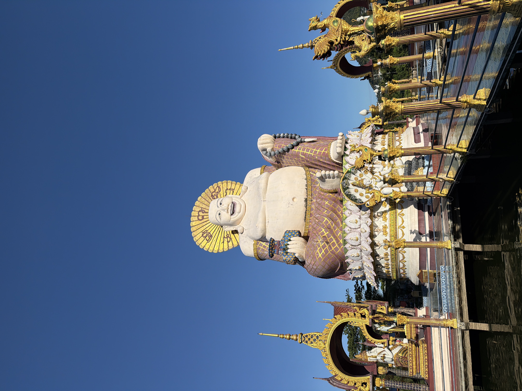The image shows a large statue of a laughing Buddha in Baan Plai Laem, Thailand. The statue is seated on a lotus flower and is adorned with a golden halo. To the left and right of the Buddha statue are ornate golden archways with mythical creatures. A walkway with golden railings leads towards the statues over a body of water. The sky is clear and blue.