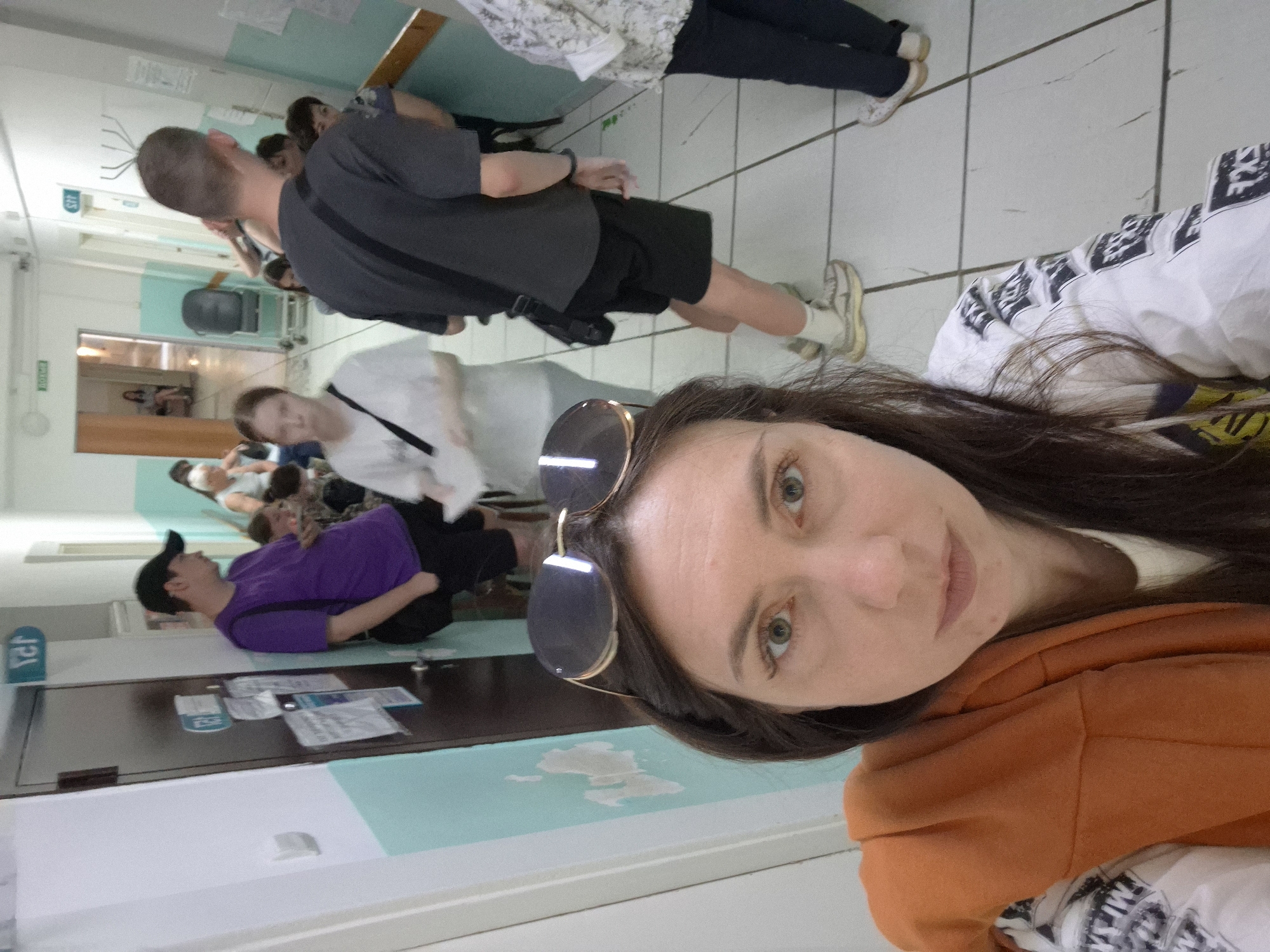 The image depicts a group of people in an indoor setting, possibly a medical facility. The woman in the foreground has long, dark hair and sunglasses perched on her head. She is wearing an orange hoodie and appears to be taking a selfie. Behind her, several individuals are visible. One man is wearing a purple shirt and black cap. Another man is dressed in a gray shirt and black shorts. People are standing and some seem to be waiting. The environment appears to be a brightly lit hallway with doors and notices on the walls. The location is Unknown, Unknown.
