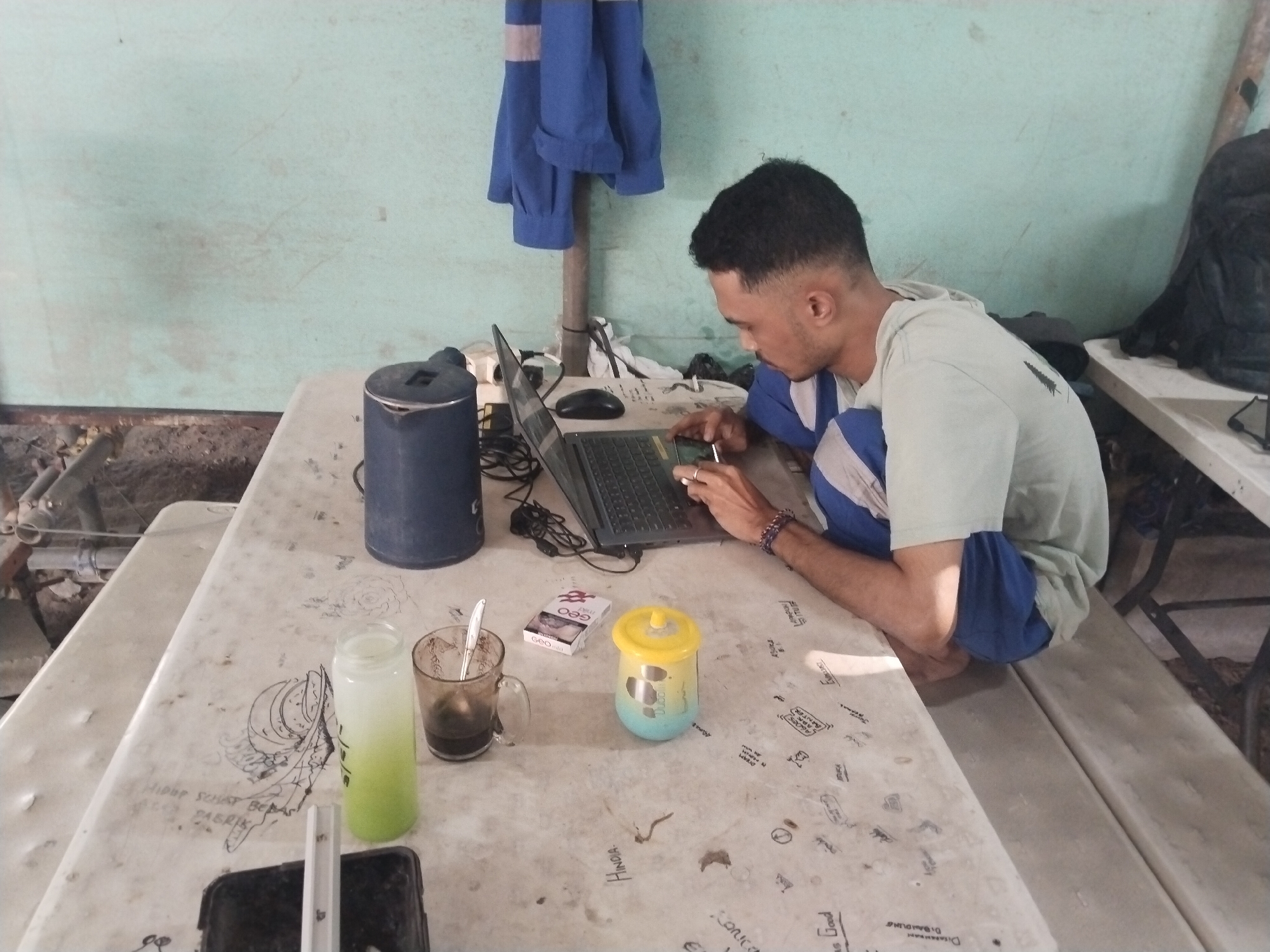 In Watuagung, Indonesia, a young man is seated at a light-colored table, working on a laptop. He is wearing a light blue shirt with white trim and blue shorts. The table is covered in various writings and drawings. On the table are a dark blue thermos, a laptop, a mouse, a cigarette pack, a glass of dark liquid with a spoon, a yellow and blue cup, and a clear bottle with a green liquid. The background shows a light green wall and a blue garment hanging on a pole. A dark backpack is visible on a bench to the right.