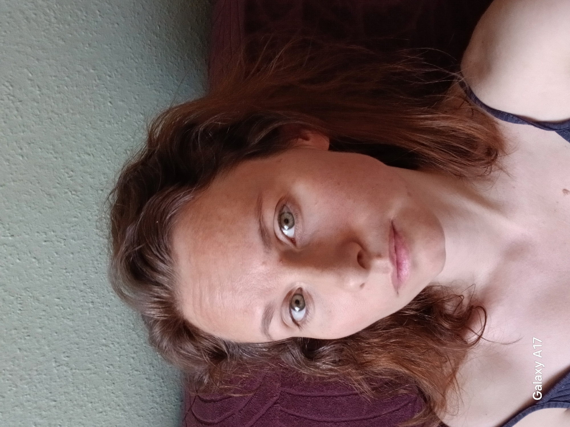 A direct portrait, possibly a selfie, features a light-skinned individual with reddish-brown, wavy hair, lying down and gazing towards the viewer. The person's head is tilted to the right, with their hair fanning out around them, resting partly on a dark red or maroon textured surface, likely a cushion or piece of furniture. Their eyes are green or hazel, reflecting ambient light, and their expression is neutral. Fine lines are visible on the forehead and around the eyes, consistent with natural skin texture.
The individual is wearing a dark strap, likely part of a top or undergarment, visible on their right shoulder and chest. The background on the left consists of a textured, muted teal or light green wall. The lighting is soft and diffused, creating a calm atmosphere indoors. There are no strong shadows, suggesting natural or evenly distributed artificial light. The image captures a close-up perspective of the person's face and upper chest.
In the lower right corner, faint white text reads "Galaxy A17," indicating the device used to capture the image. The scene is set in Yekaterinburg, Russia, although no specific external location cues are visible within the image itself.